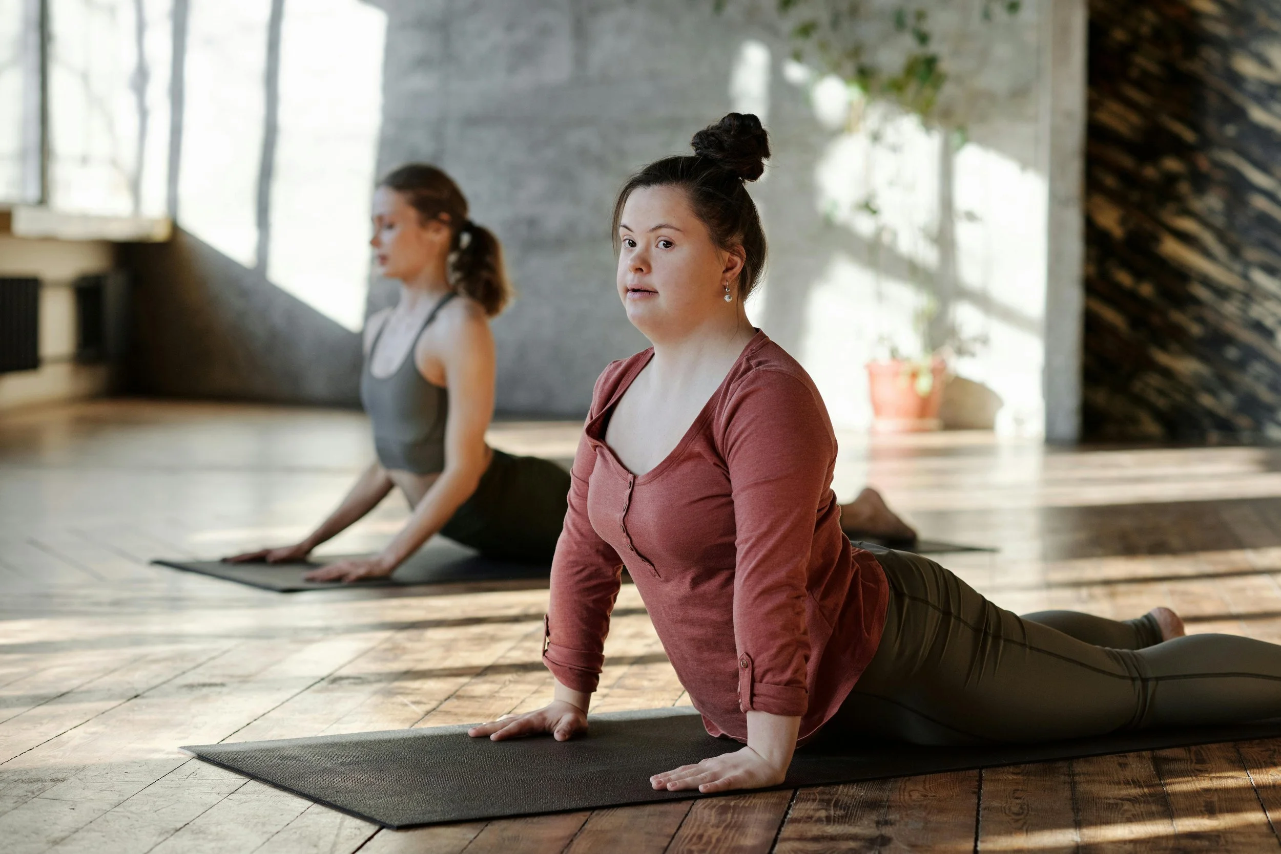 Two women practicing yoga on mats in a studio, one in the foreground and one in the background, both in a kneeling pose with hands on the floor.