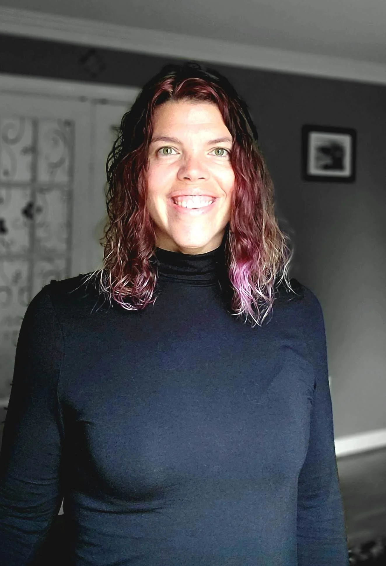 A woman with curly hair that is dark with purple and pink highlights, wearing a black turtleneck, smiling indoors.