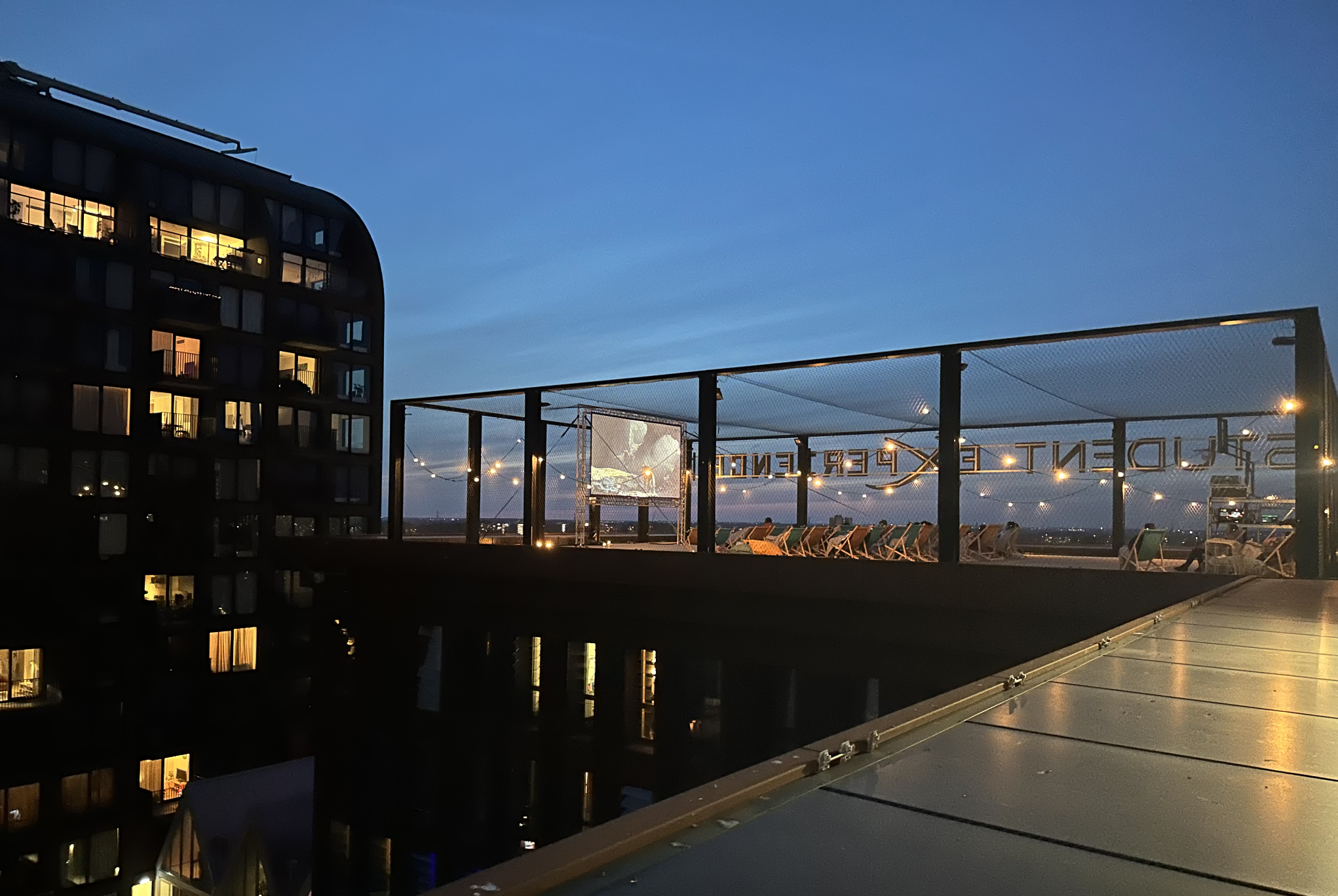 Rooftop with lounge chairs and string lights at dusk, overlooking city skyline.