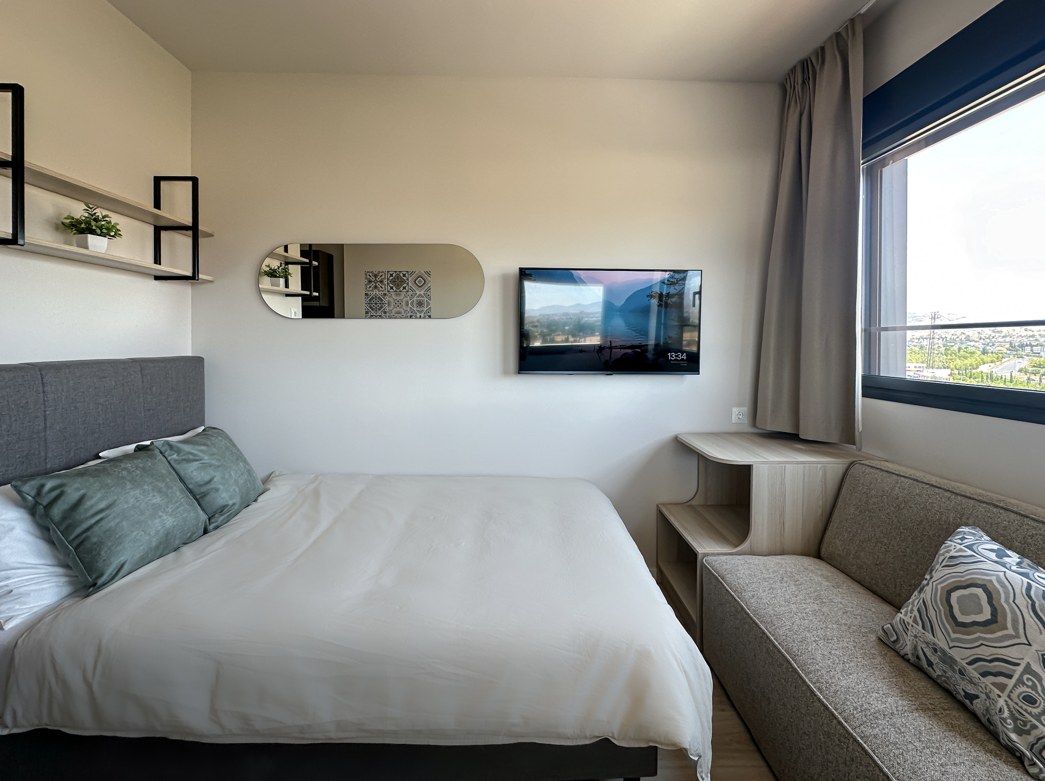 Hotel bedroom with a bed, wall-mounted flat-screen TV, window with view, mirror, and seating area.