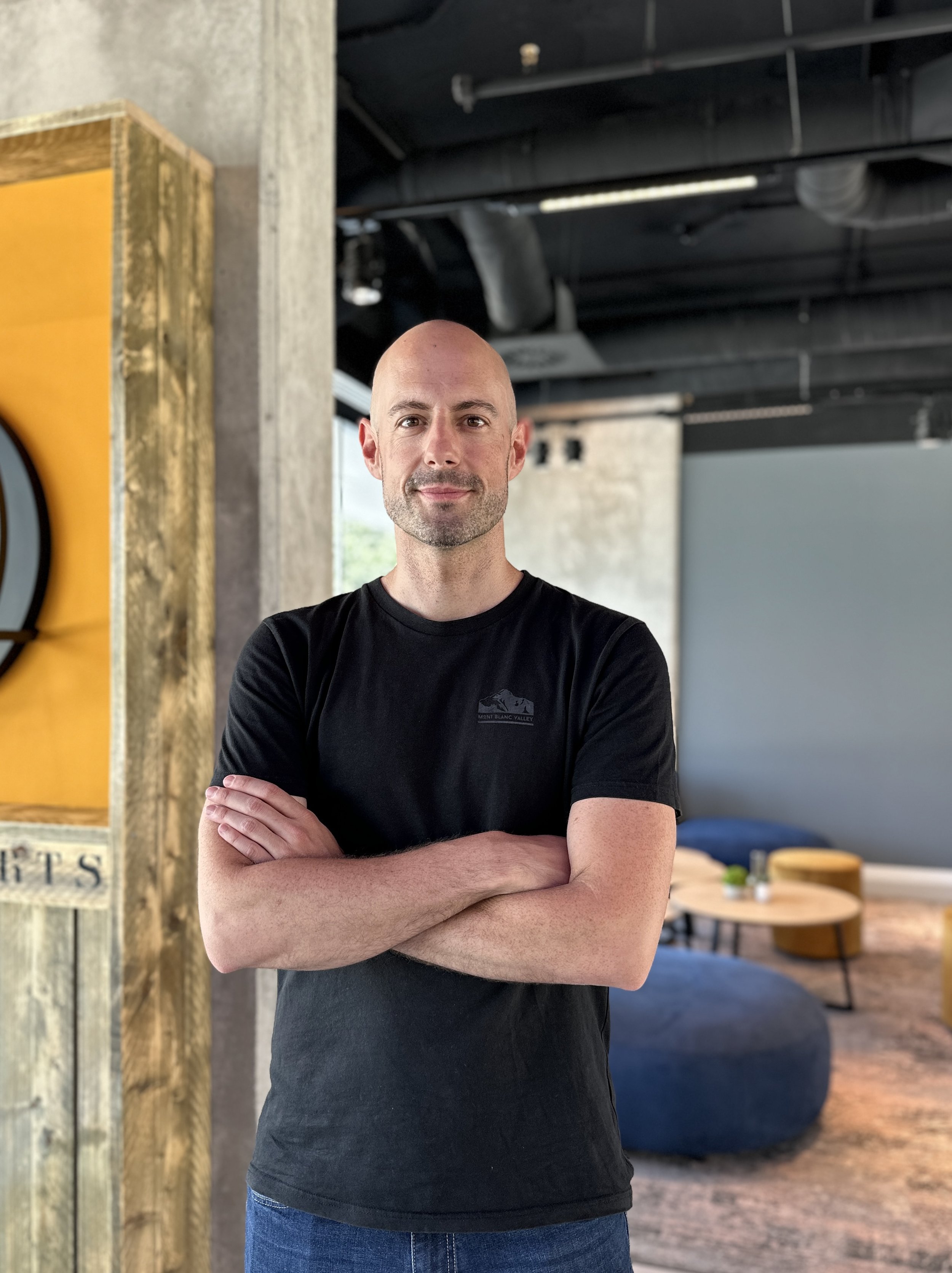 A man standing with arms crossed in a modern industrial-style room, wearing a black t-shirt, with a neutral background and seating area behind him.