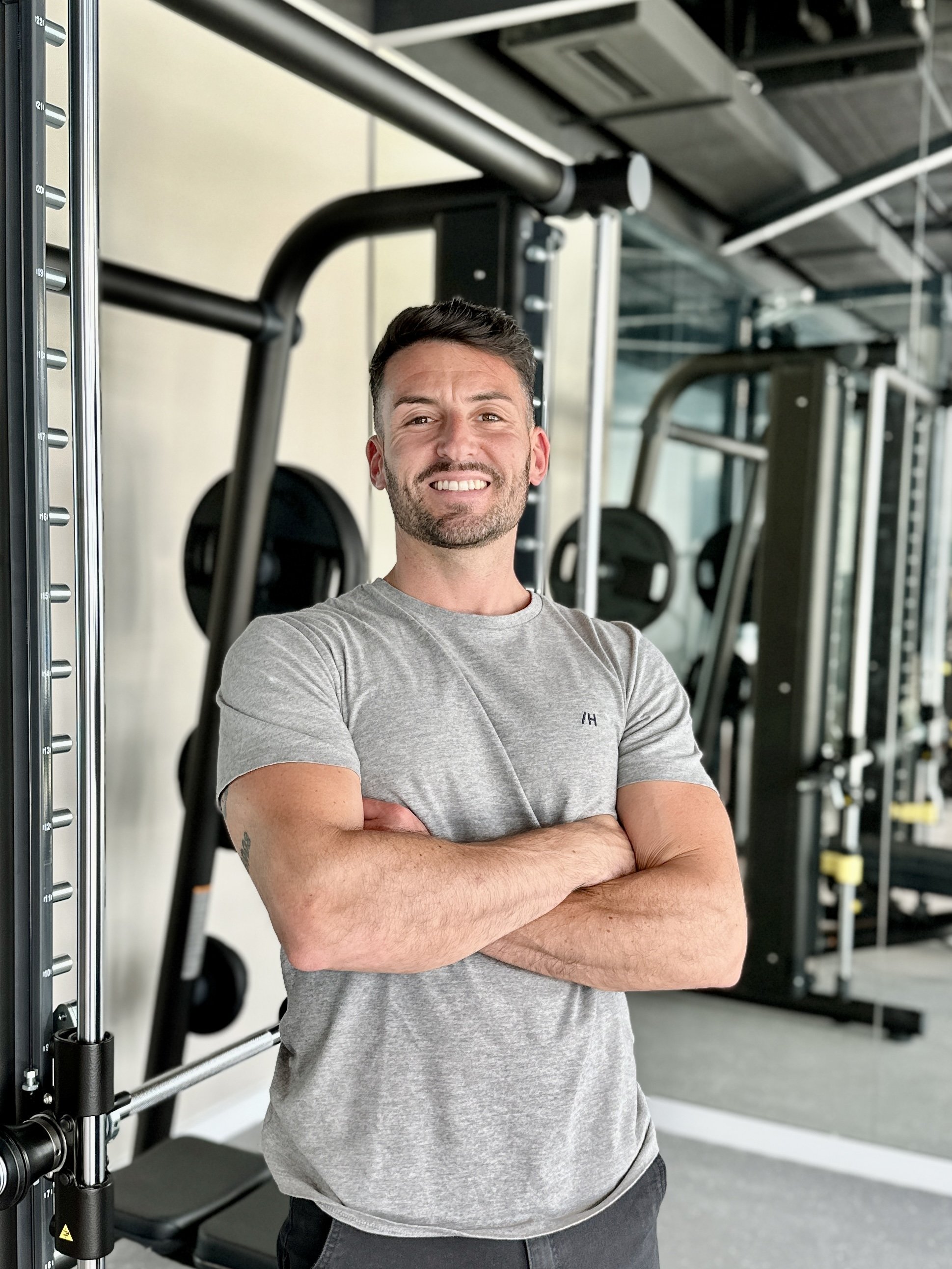 A young man with a beard and short dark hair standing with crossed arms in a gym, smiling at the camera. He is wearing a light gray t-shirt, and there are weightlifting machines behind him.