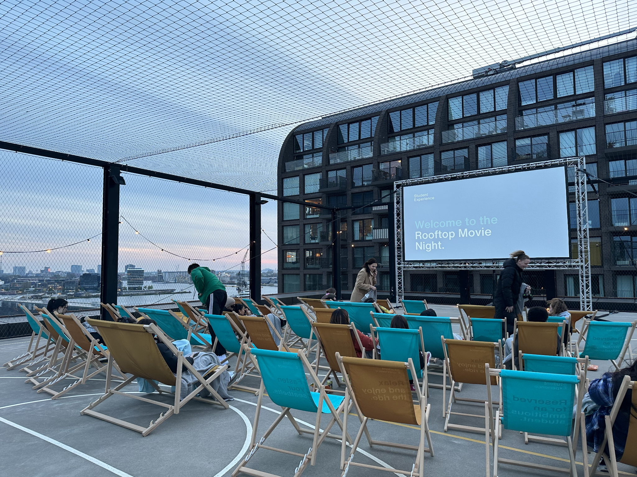 Rooftop movie night setup with chairs, a large screen displaying welcome message, and city skyline in the background during evening.
