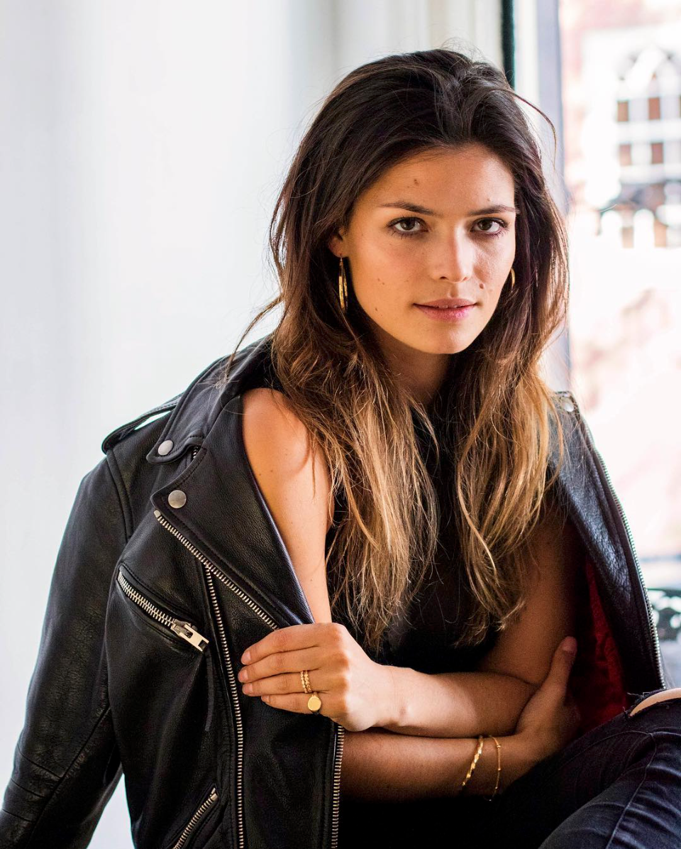 A woman with long brown hair, wearing a black leather jacket, gold jewelry, and black top, sitting indoors near a window.