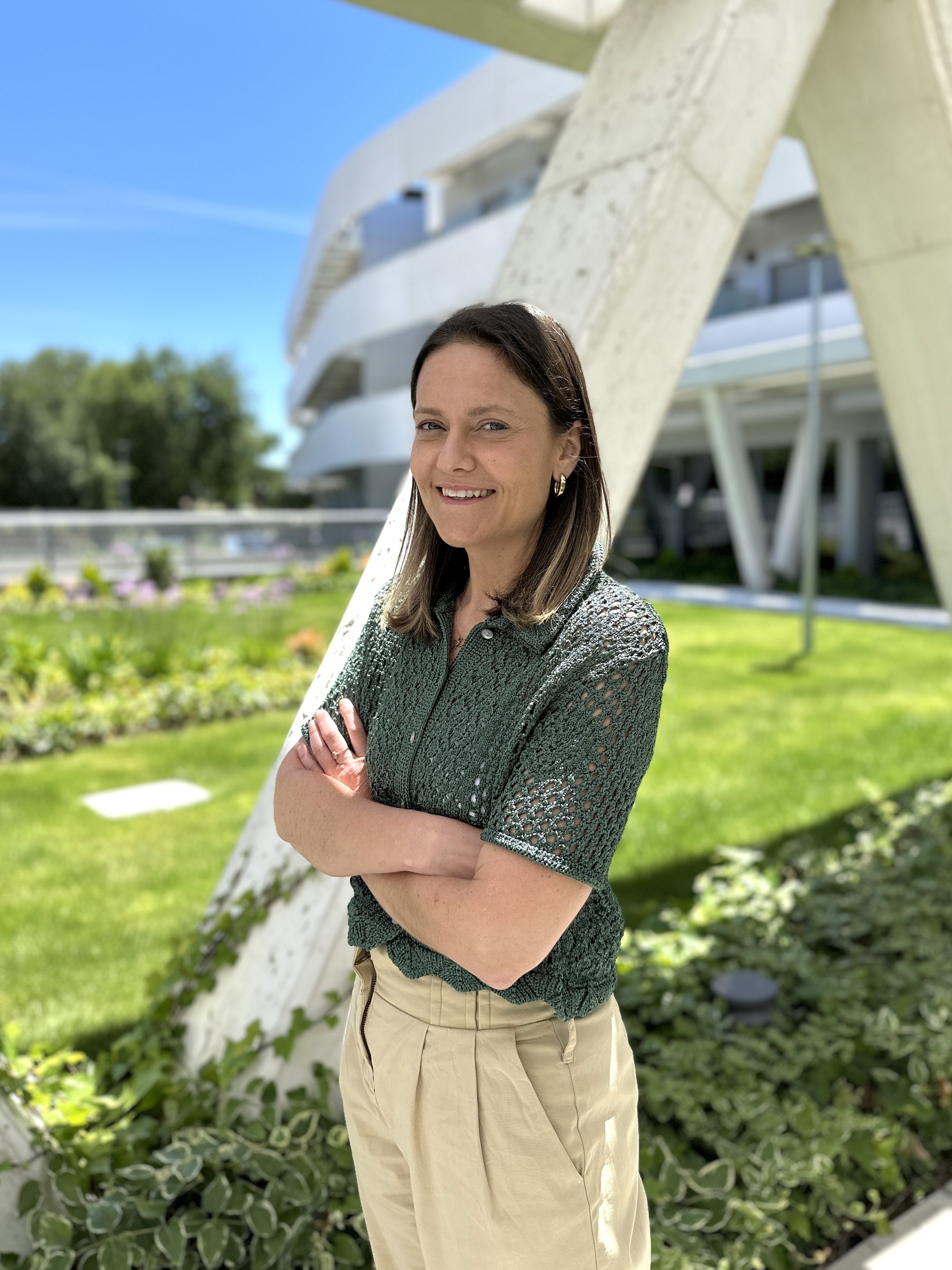 A woman with shoulder-length brown hair, wearing a green open-knit top and beige pants, stands outdoors in front of a large white modern architectural structure, smiling with arms crossed, on a sunny day.