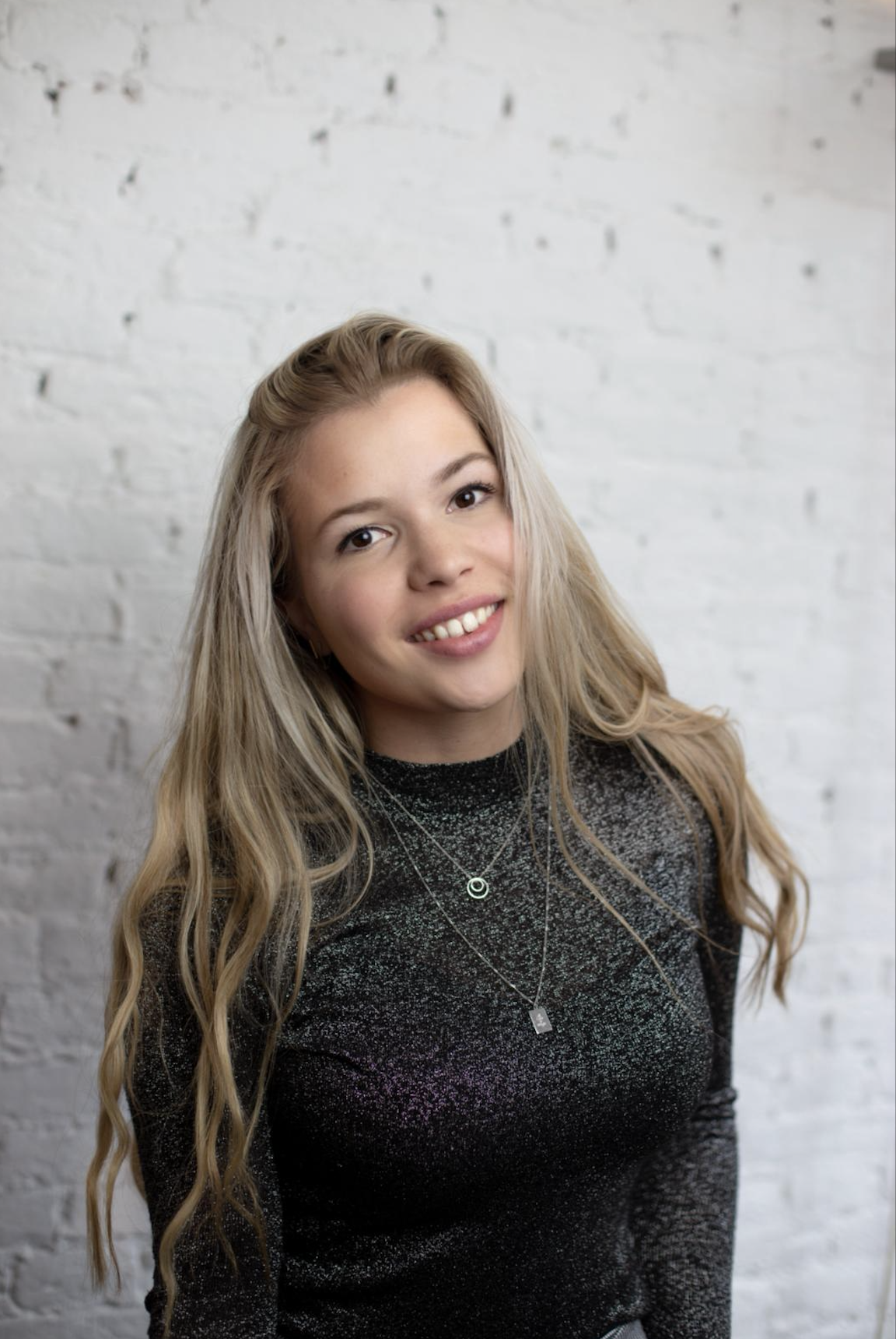A young woman with long wavy blond hair smiling, wearing a black sparkly top and layered necklaces, standing in front of a white brick wall.