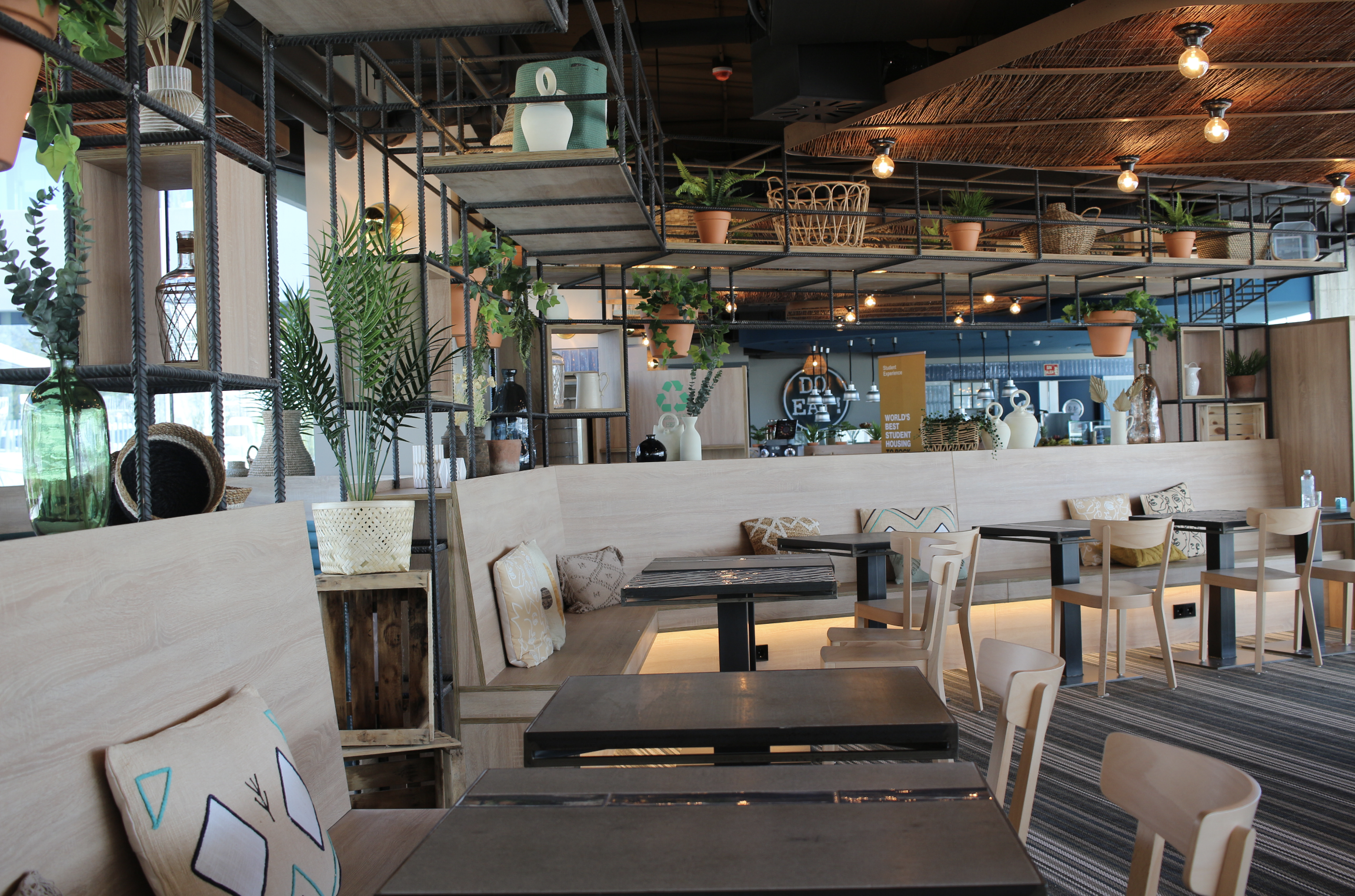 Modern restaurant interior with wooden furniture, decorative cushions, potted plants, and open shelving with vases and decorative items, illuminated with warm lighting.