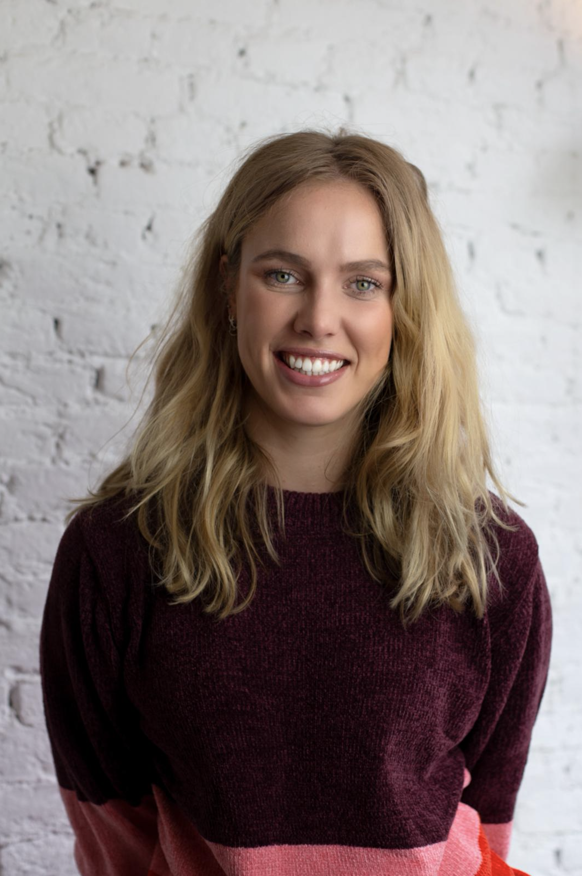 A smiling woman with blonde hair, wearing a maroon sweater, standing in front of a white brick wall.