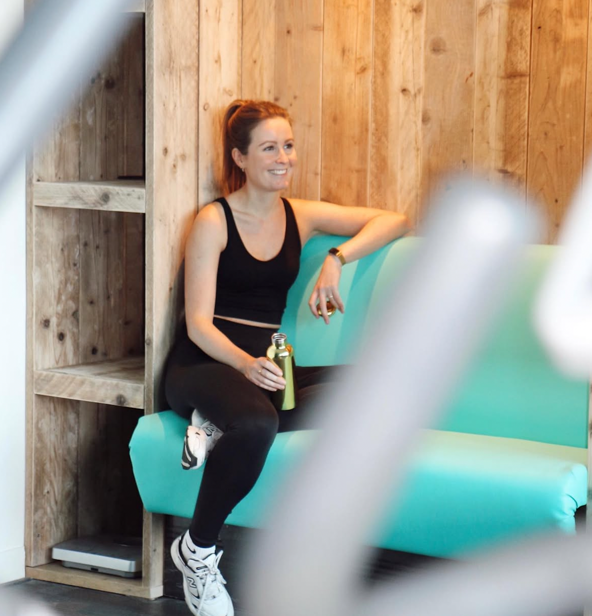 A woman with a ponytail, wearing a black sports bra and black leggings, sitting on a green couch in a wooden room, smiling and holding a metal water bottle.