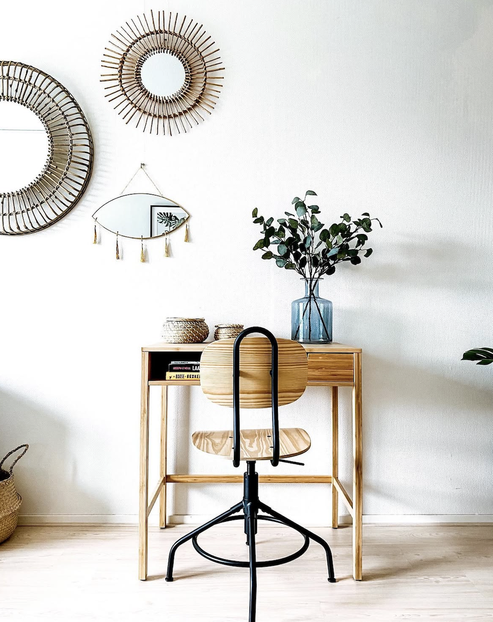 A minimalist home office setup with a wooden desk, ergonomic wooden chair, vase with green foliage, decorative wall mirrors, and a mirror hanging on the wall, all against a white wall and light wooden flooring.
