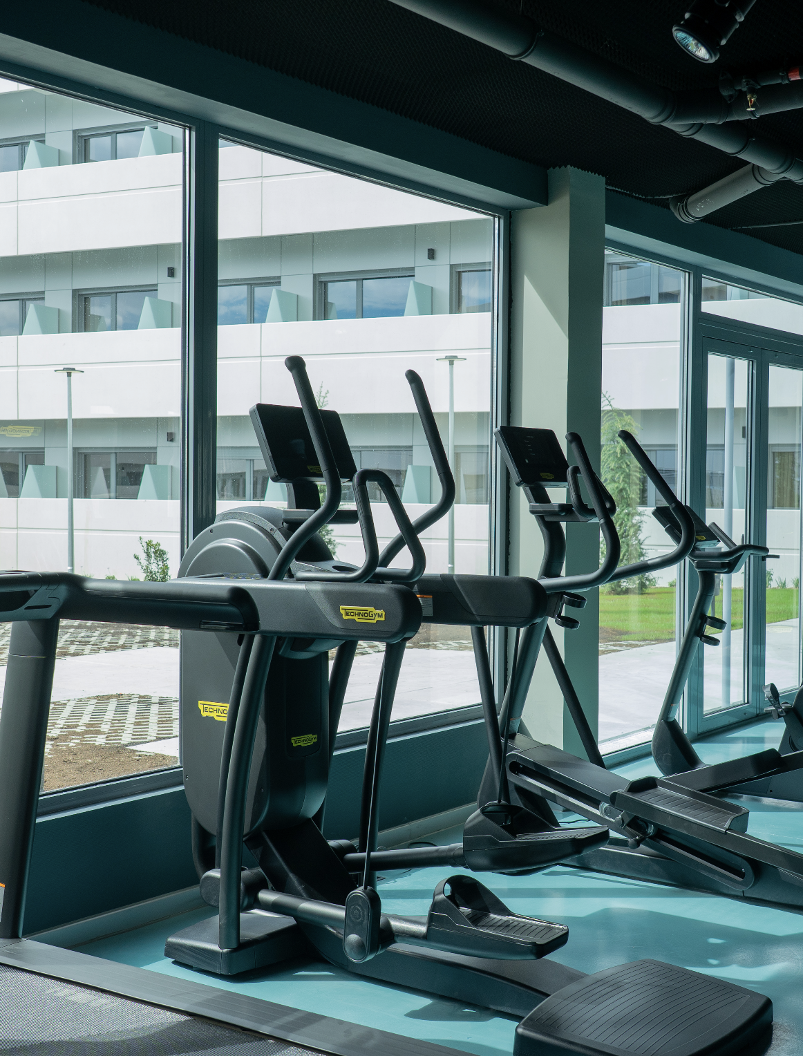 Gym with exercise machines including ellipticals near large windows with a view of an apartment building and outdoor greenery.