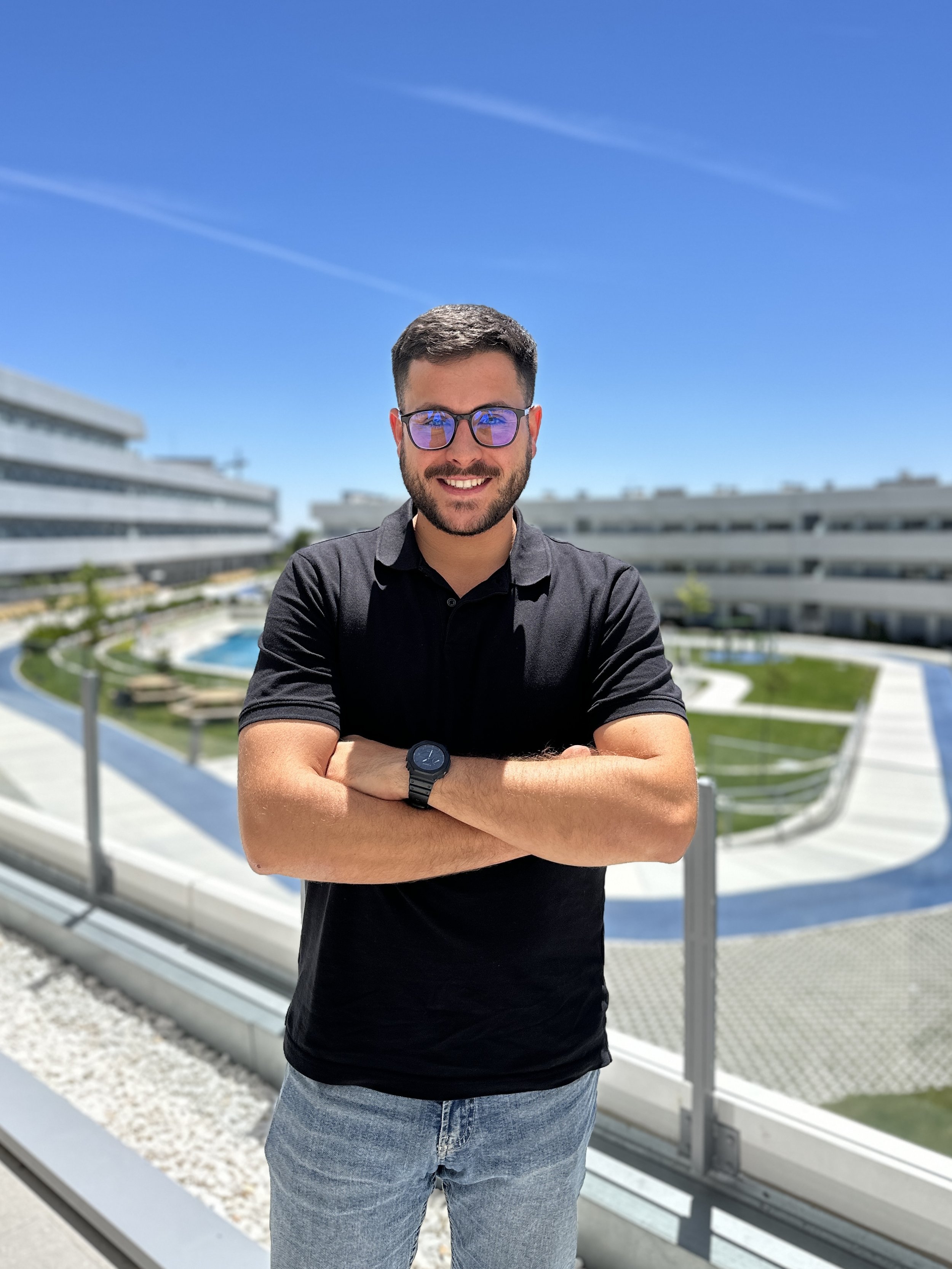 Young man with glasses, wearing a black polo shirt and jeans, standing outdoors with folded arms, smiling, in front of a modern residential complex with a courtyard and swimming pool under a clear blue sky.