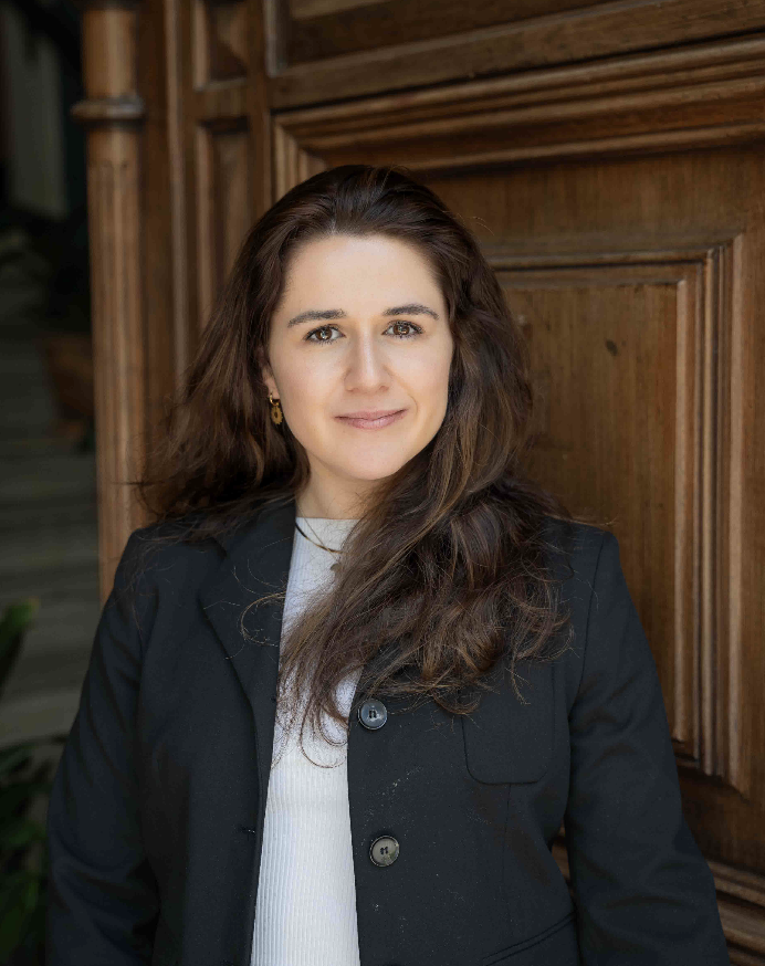 A woman with shoulder-length brown hair and light skin, wearing a black blazer over a white top, standing indoors against a wooden wall and looking at the camera with a slight smile.