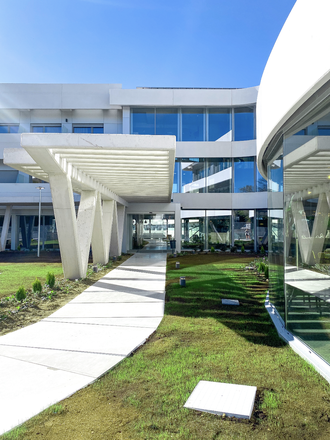 Modern building with glass windows and white concrete structure, curved glass wall on the right, green lawn and pathway leading to entrance under a blue sky.