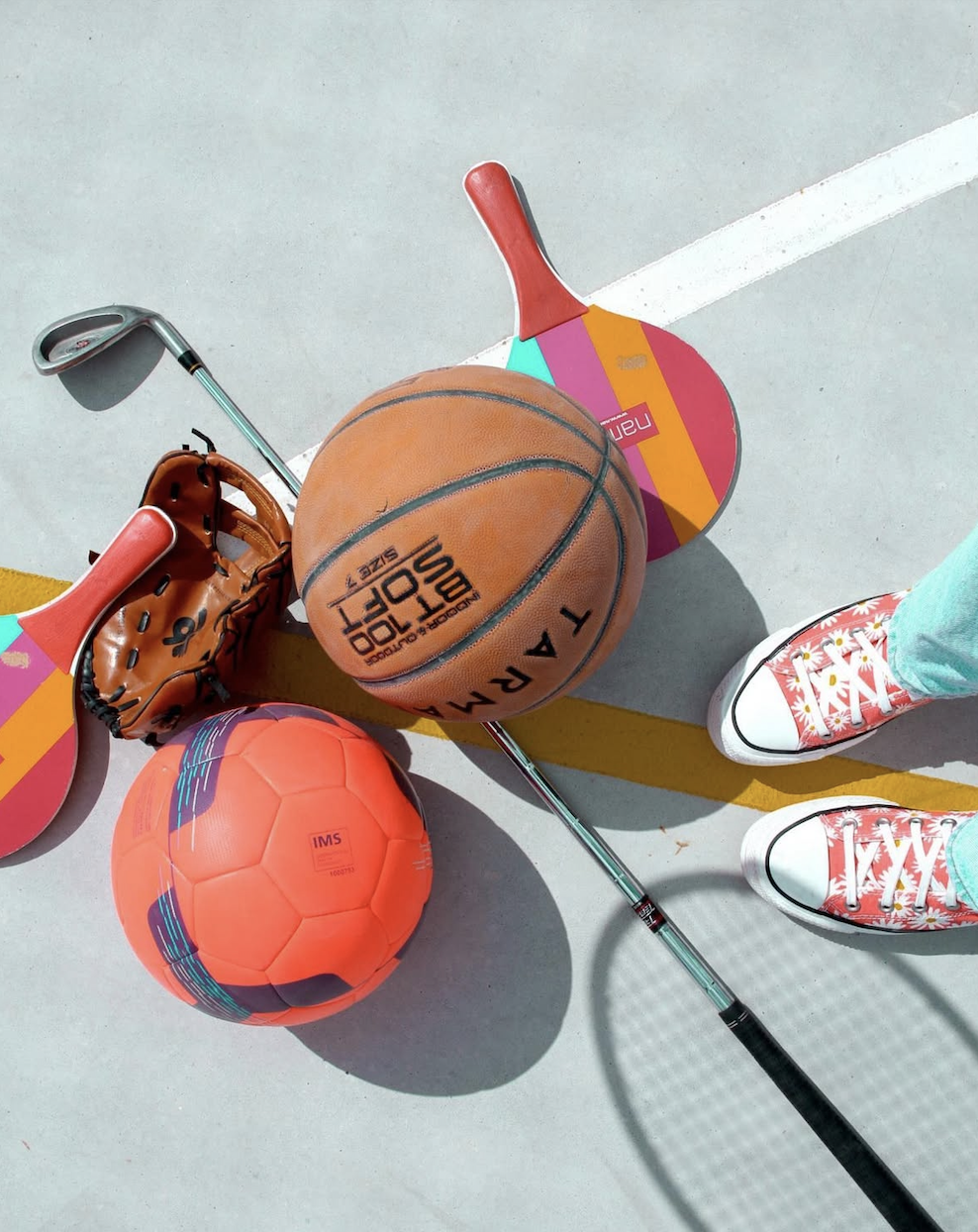 Basketball, tennis racket, pickleball paddle, soccer ball, baseball glove, and a person's feet in colorful sneakers on a tennis court.