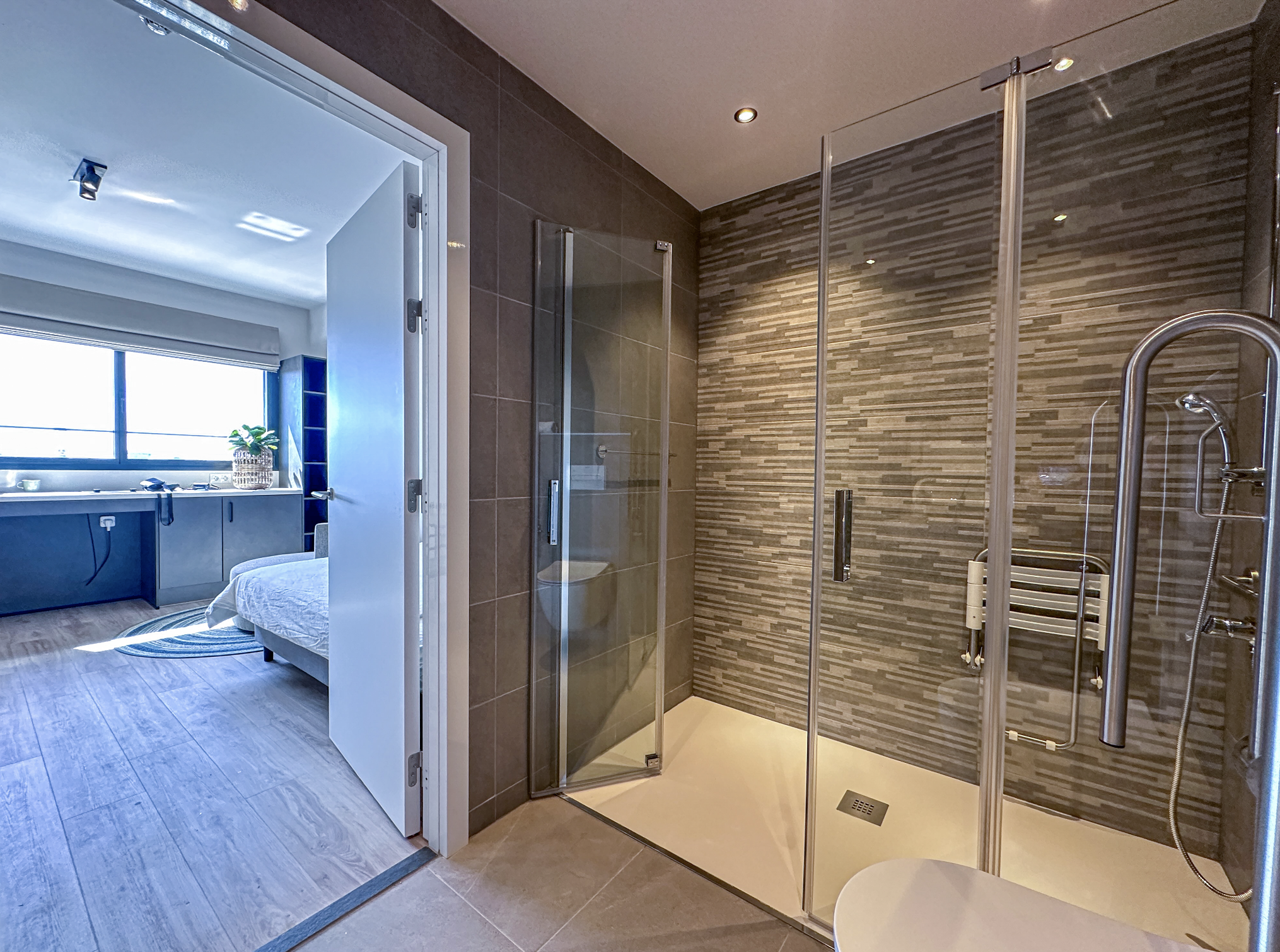 Modern bathroom with a walk-in shower featuring dark textured wall tiles, a glass door, and a shower head, opening into a bedroom with a large window, a bed, and a potted plant.