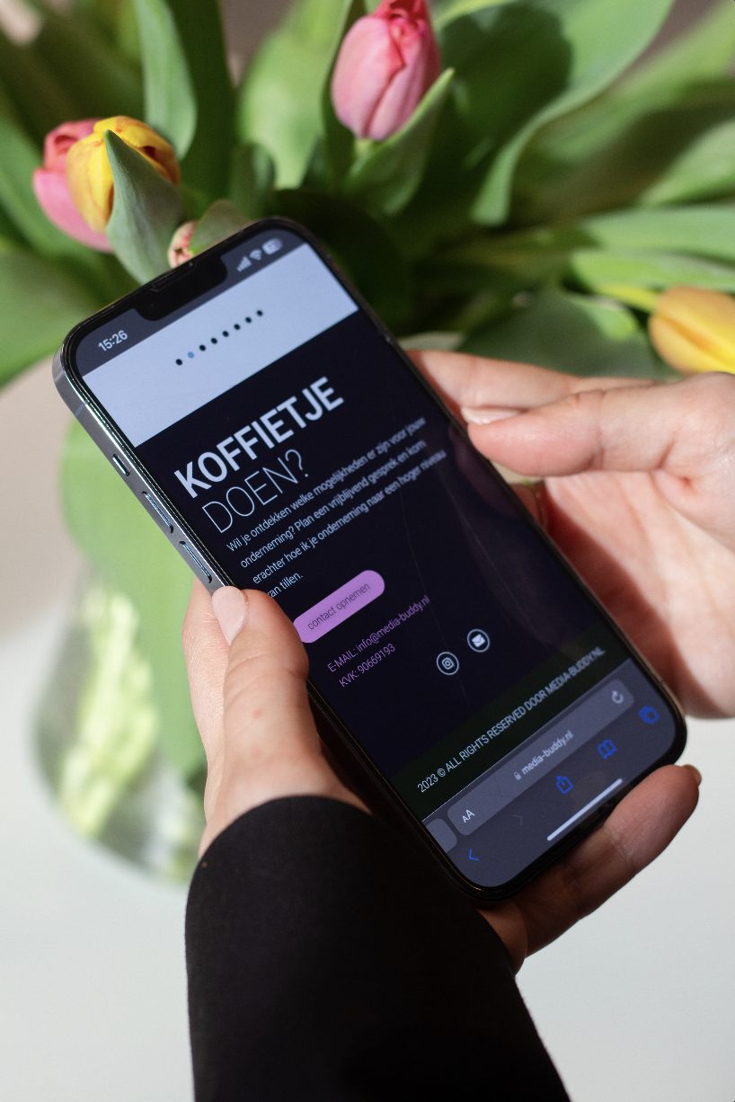 Person holding a smartphone with a website displayed in Dutch, in front of a vase of tulips.