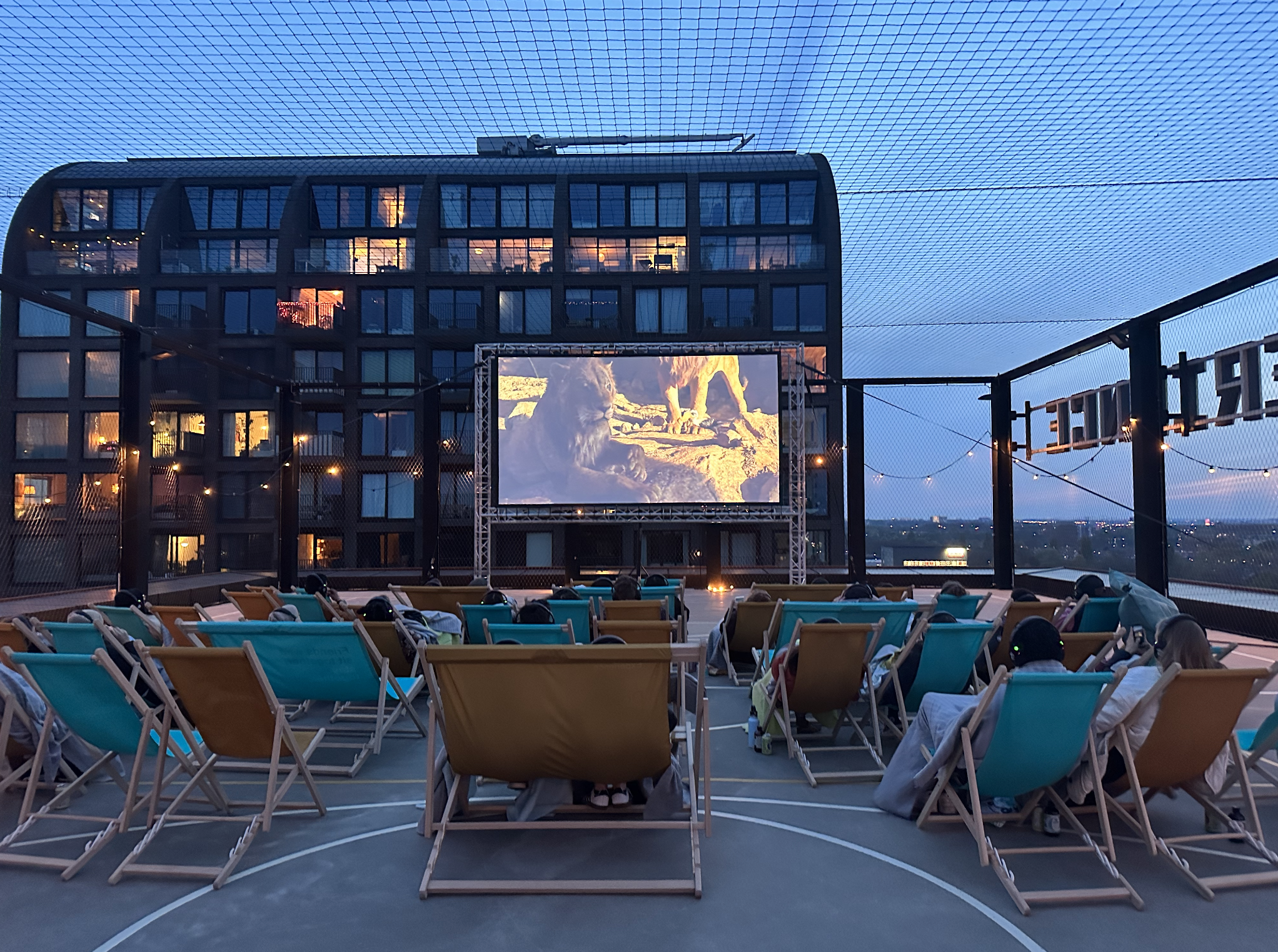 People watching an outdoor movie screening on a rooftop at dusk, with a large screen showing lions. The area has lounge chairs, a netted roof, and a city view in the background.
