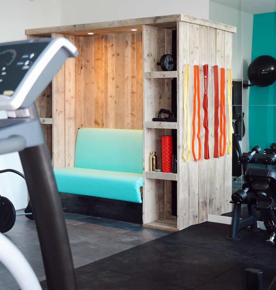 Workout area with a wooden cubicle that includes a turquoise bench, fitness equipment, resistance bands hanging, and exercise balls in the background.