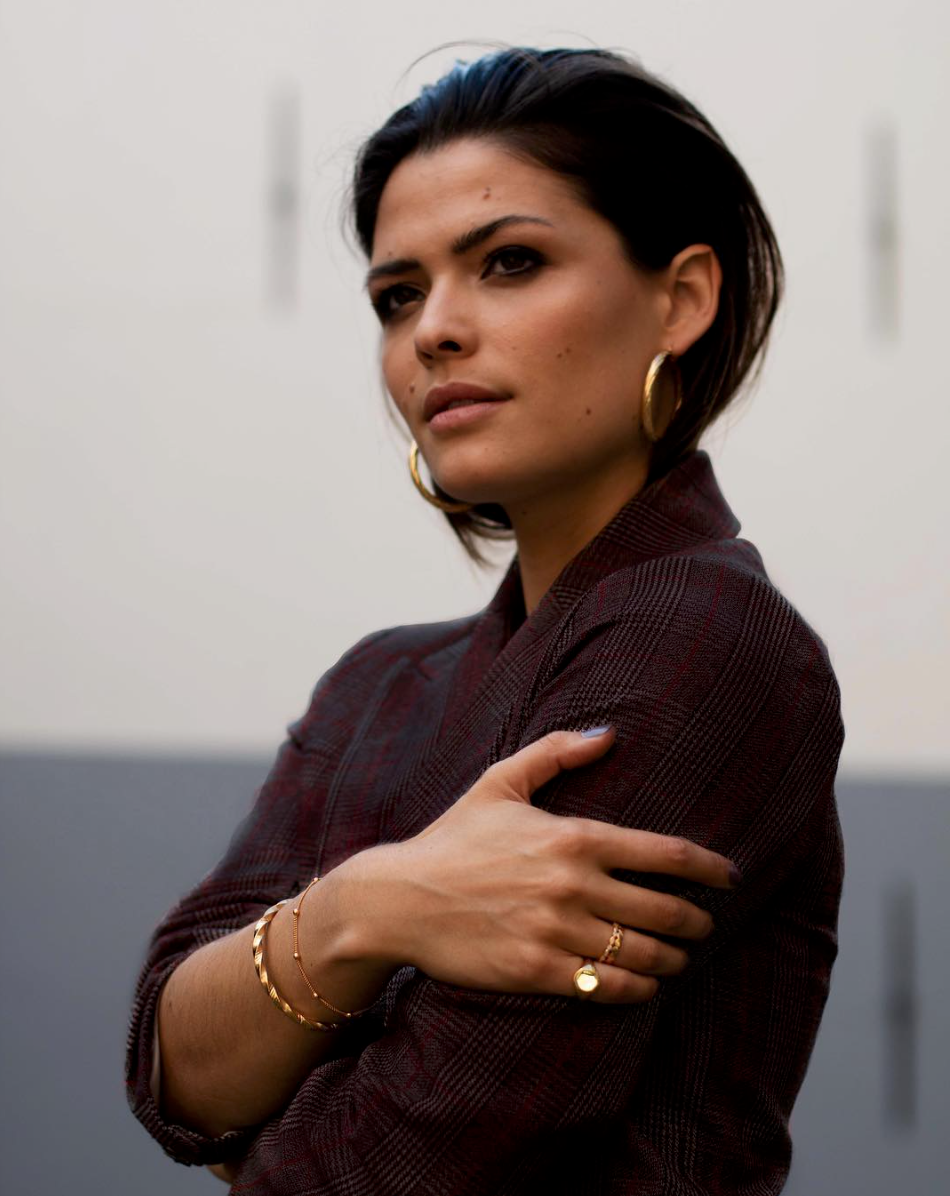 A woman with dark hair, wearing large gold hoop earrings and gold jewelry, posing with her arms crossed in front of her chest, dressed in a dark, patterned blazer, against a plain background.