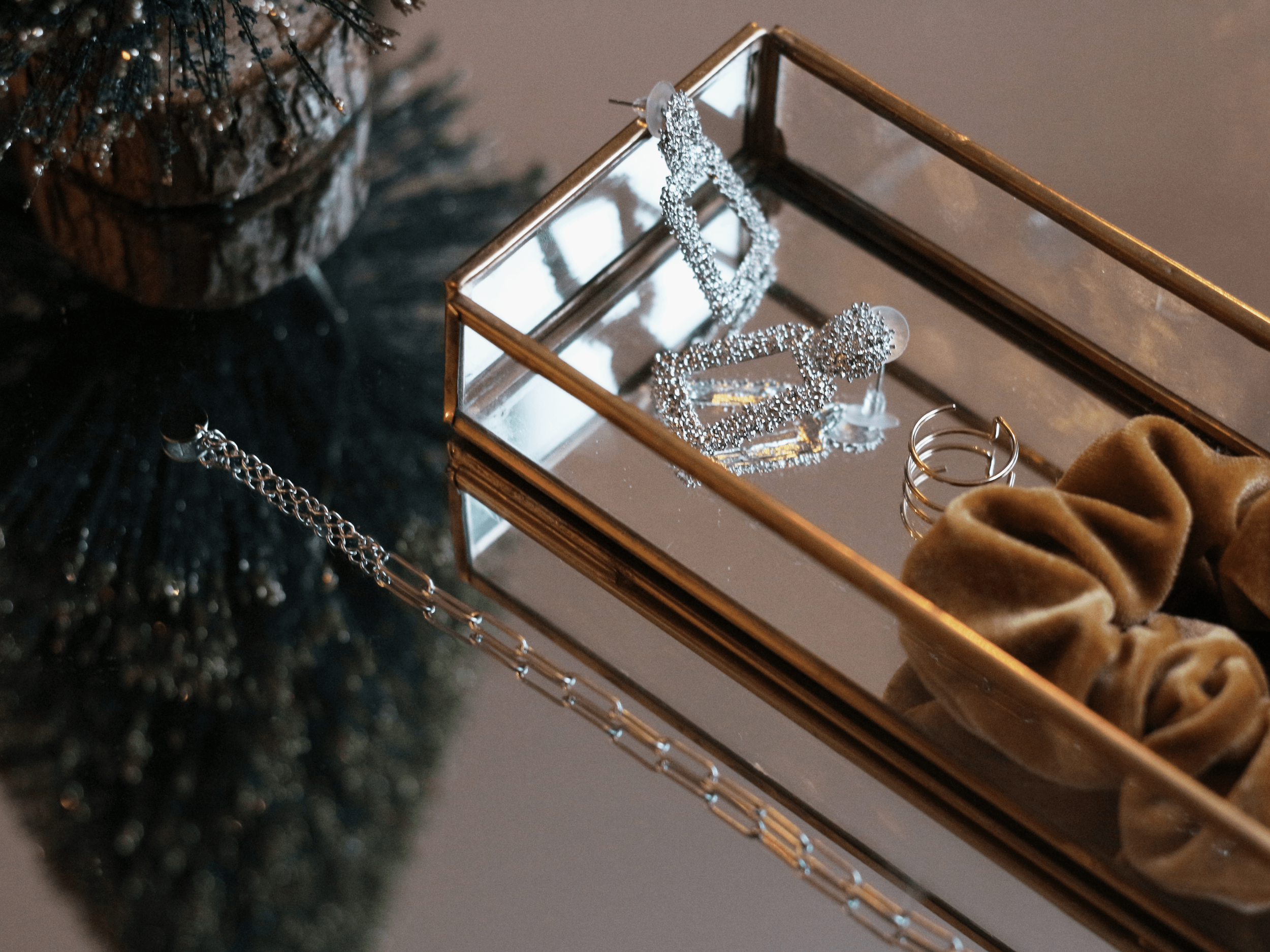 A glass jewelry box with a silver mirror base, containing a sparkly silver necklace and rings, next to a brown velvet scrunchie and a pile of dark-colored Christmas tree branches.