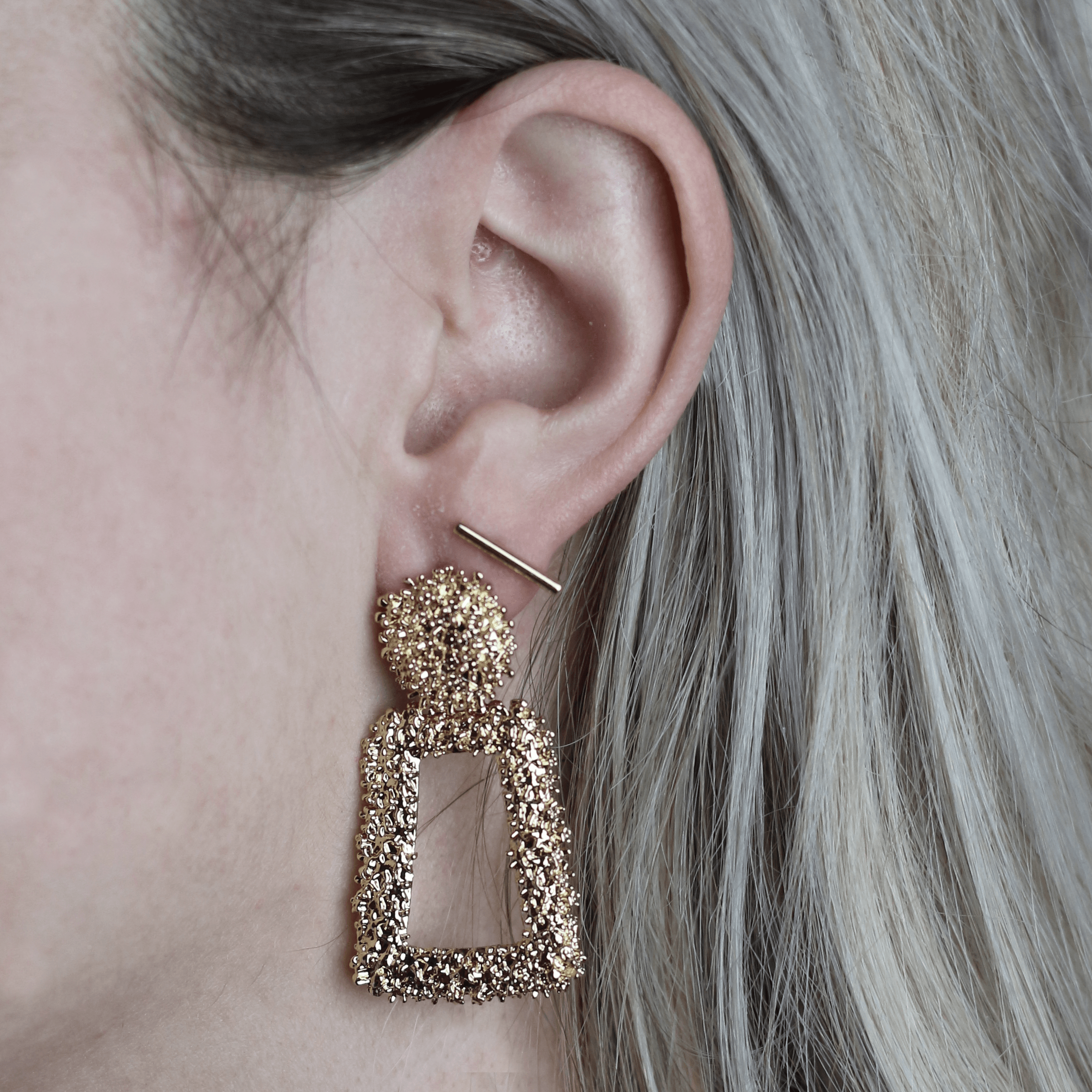 Close-up of a person's ear wearing a gold, textured, geometric earring and a small gold bar earring.