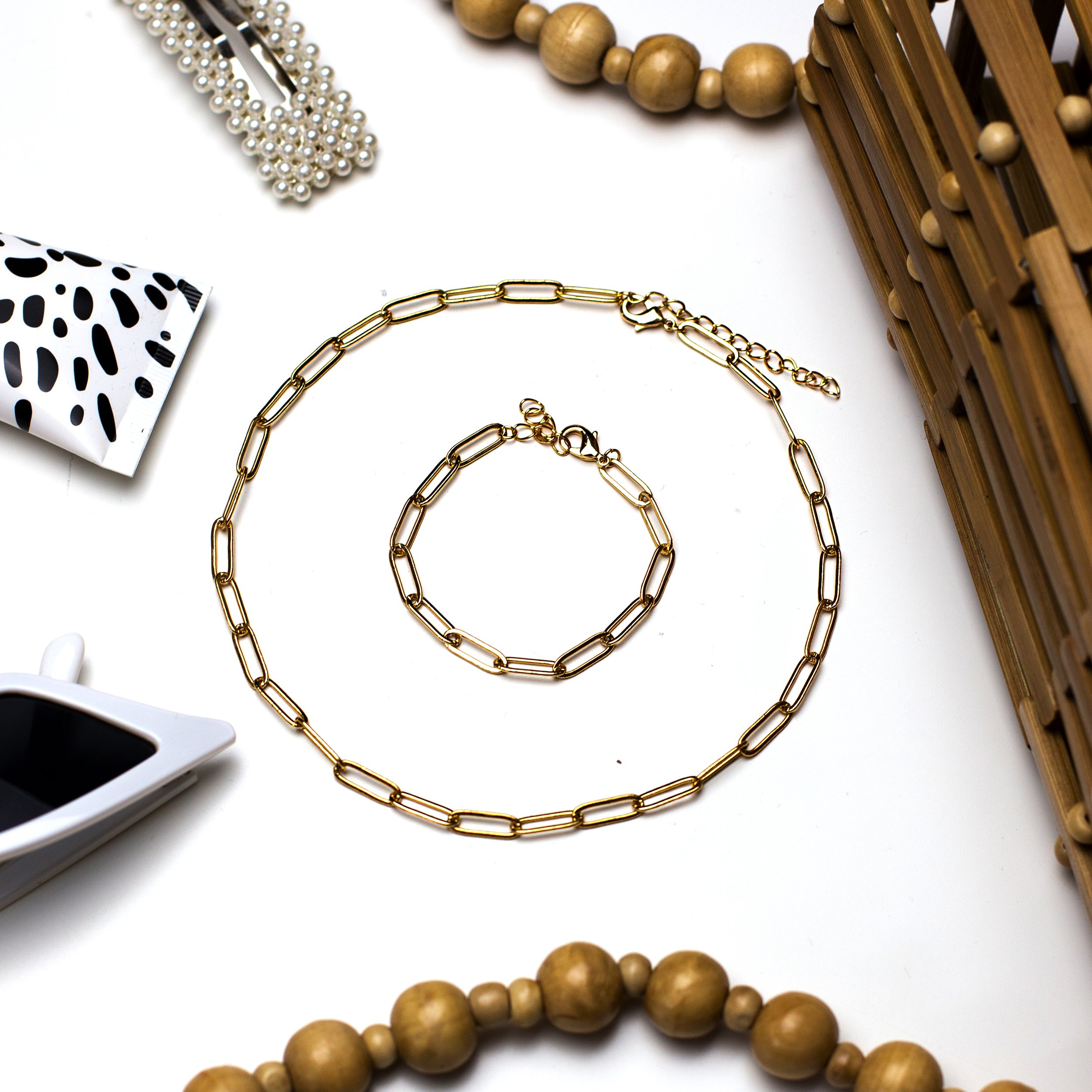 Gold chain necklace and matching bracelet on a white surface, surrounded by pearl jewelry, a wicker basket, a white coffee cup with black design, and a black-and-white patterned item.