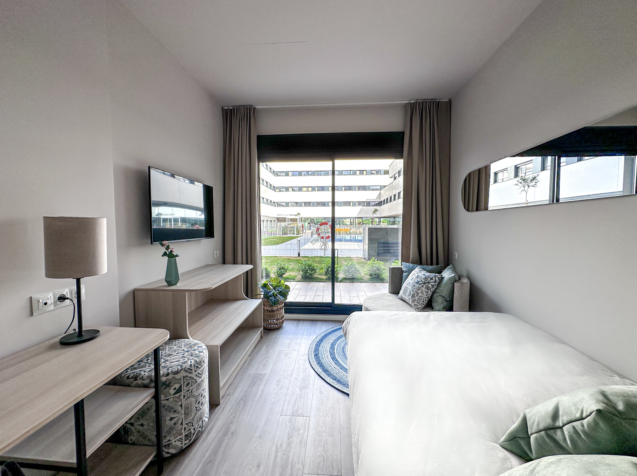 Modern hotel room with a large bed, a small sitting area with a sofa and pillows, a flat-screen TV mounted on the wall, a wooden desk with a lamp and patterned stool, and a sliding glass door leading to a balcony with a view of other modern buildings