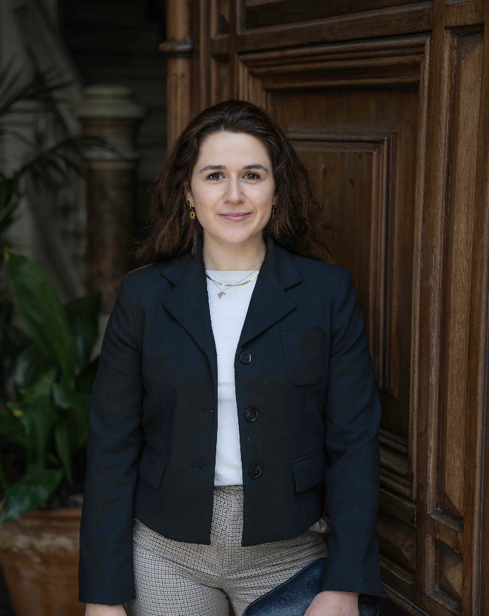 A woman with long dark hair wearing a navy blazer, white top, and checkered pants, standing indoors near wooden paneling and green plants.