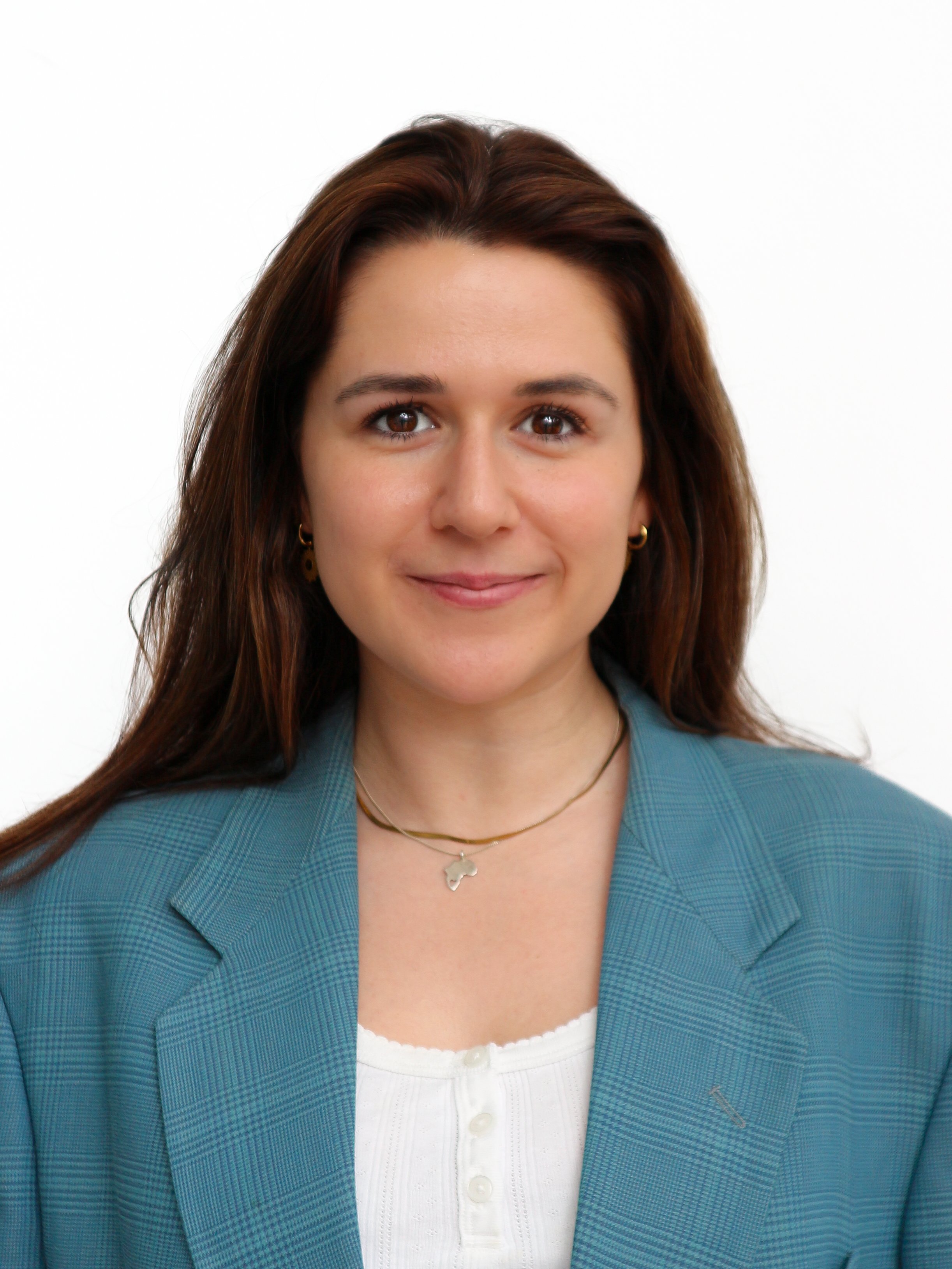 Headshot of a woman with long, brown hair, wearing a teal blazer over a white top, small gold earrings, a gold necklace with a star-shaped pendant, and smiling against a white background.