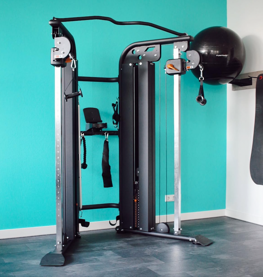Multifunctional gym machine with a black exercise ball mounted on the right side, set against a teal wall in a fitness room.