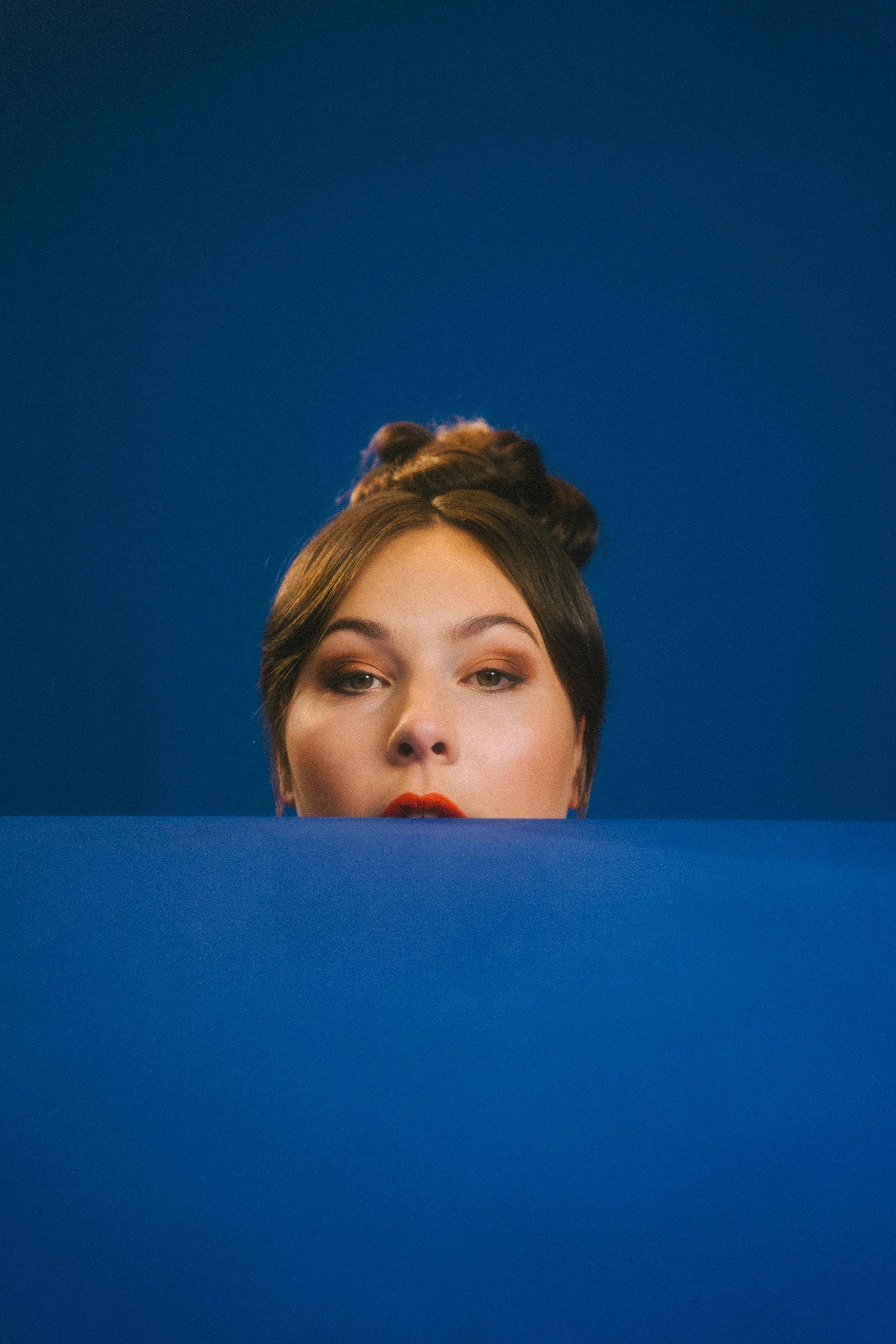 Eine Frau mit Hochsteckfrisur und roten Lippen schaut über eine blaue Wand, vor blauem Hintergrund hervor.