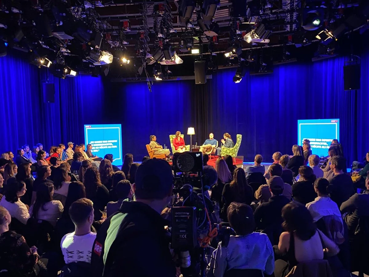 Bühnenveranstaltung mit Publikum und Moderatorengruppe, von Kamera und Beleuchtung umgeben, in einem Studio mit blauer Beleuchtung.