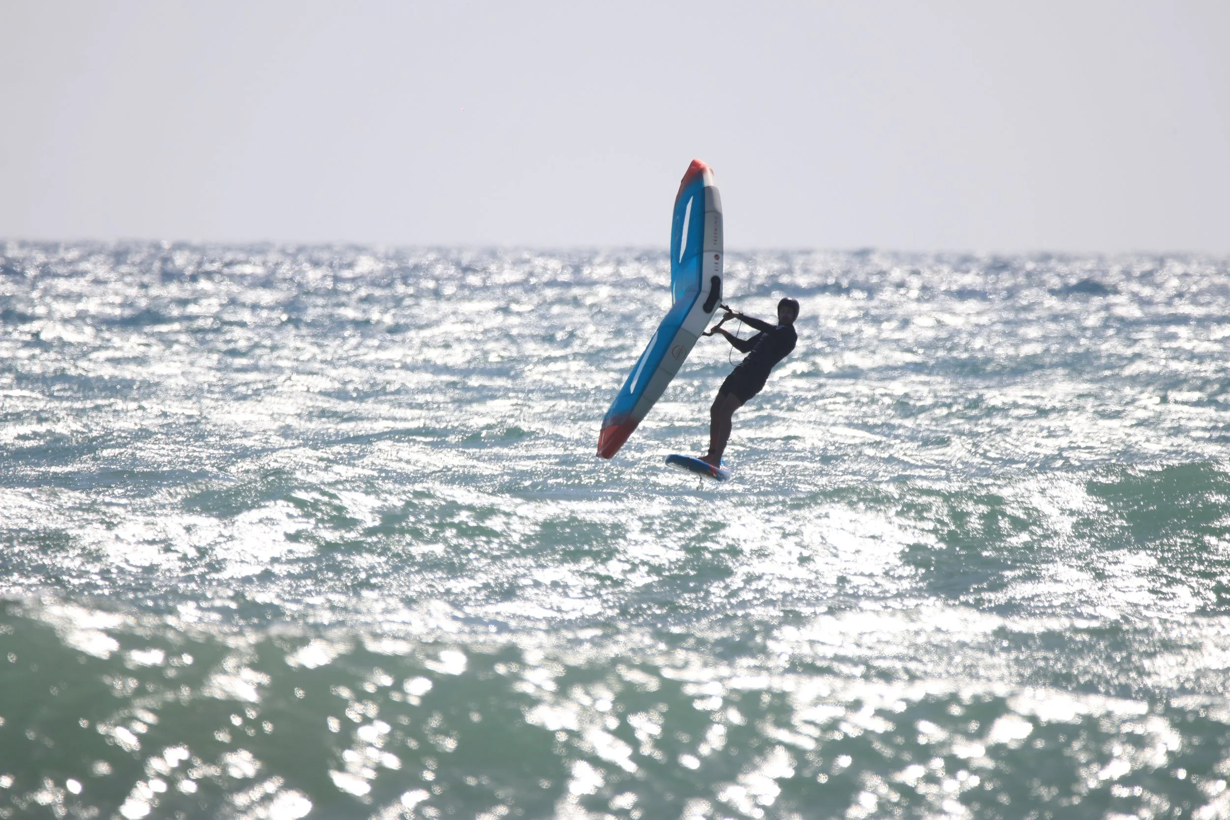 Person windsurfing on the ocean, holding onto the sail, with the water sparkling under sunlight.