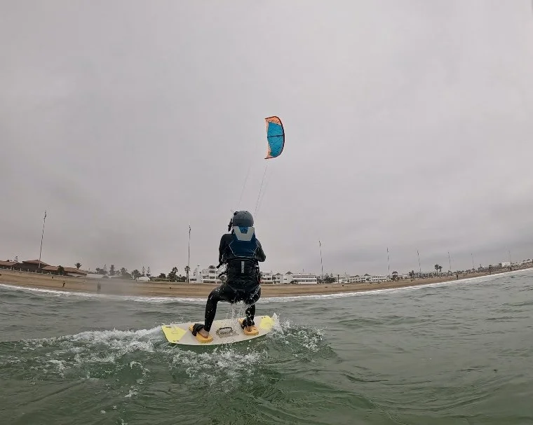 kitesurf student with helmet kiteboarding on the ocean with a colorful kite, buildings and palm trees visible on the shoreline in the background under cloudy sky.