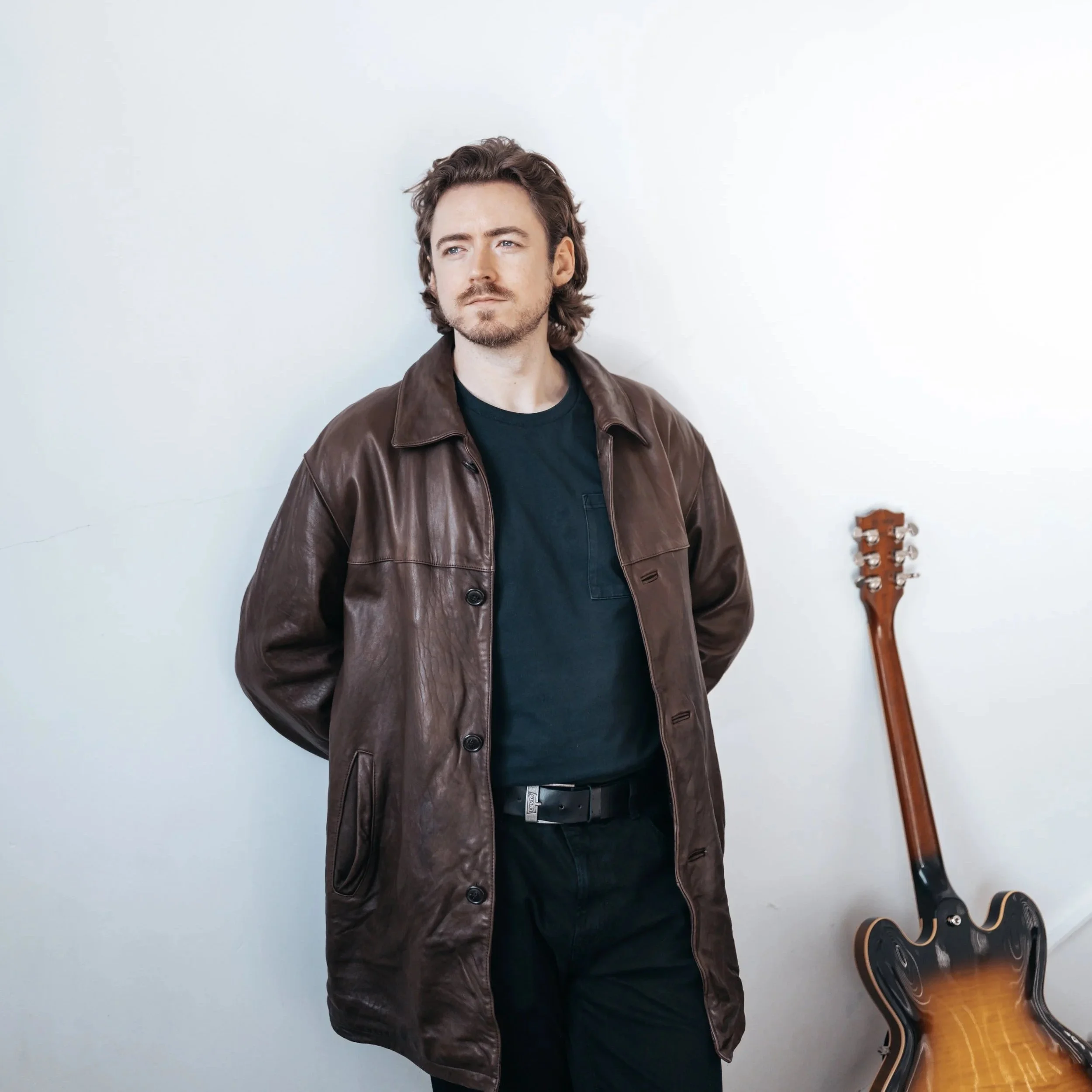 A man in a brown leather jacket stands against a plain white wall, with a guitar resting on the floor beside him.