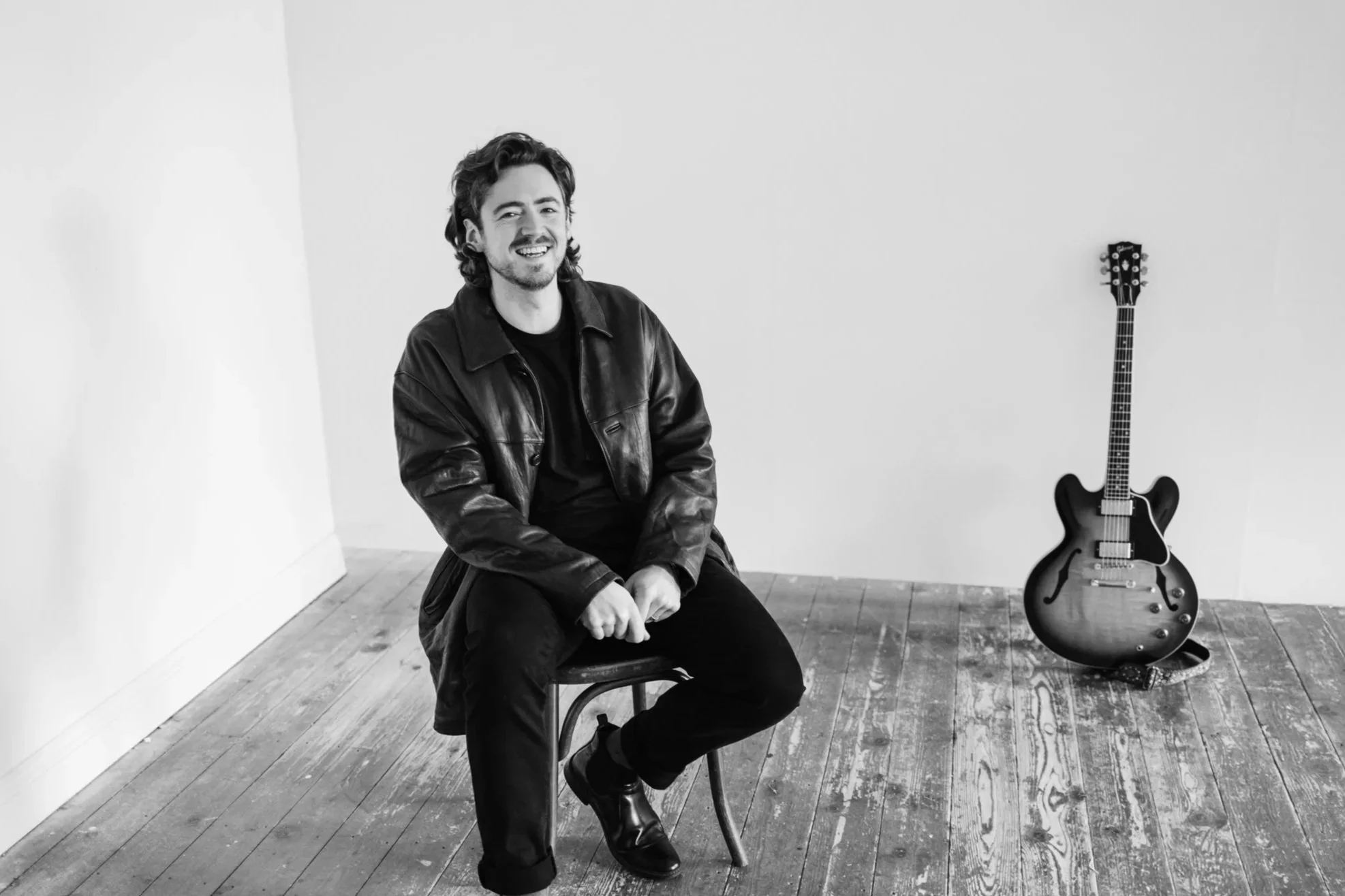A man sitting on a chair in a minimalistic room with a guitar against the wall, all in black and white.