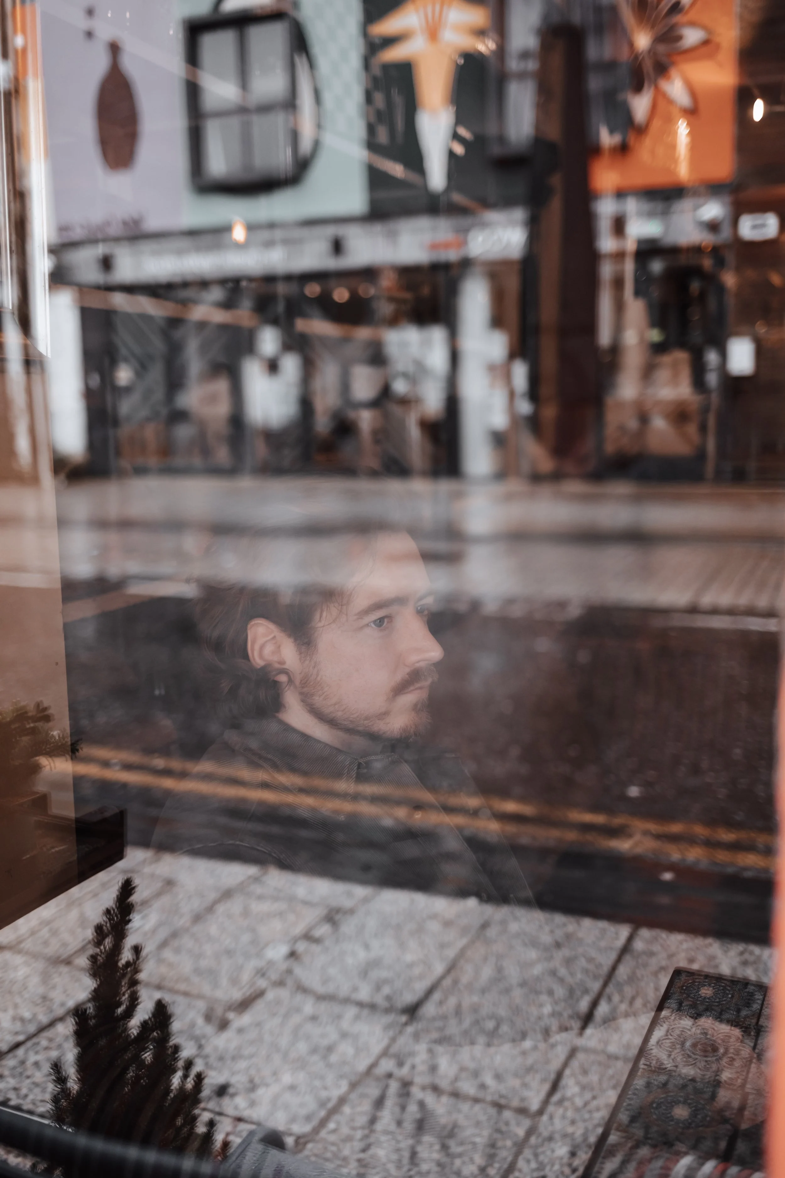 A reflection of a man sitting inside a cafe looking out the window at the street with cobblestone sidewalk. The cafe interior features framed art, plants, and warm lighting.