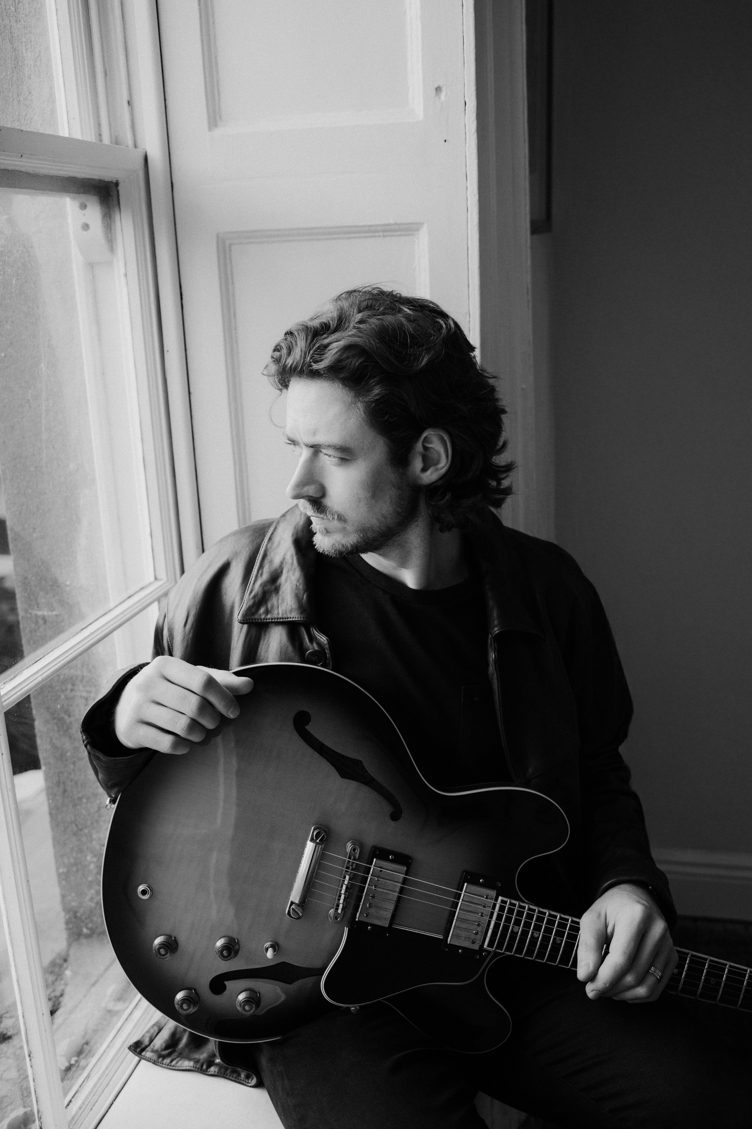 A young man with wavy hair and a beard, wearing a leather jacket, sitting by a window holding a semi-hollow electric guitar, looking out contemplatively in a black-and-white photo.