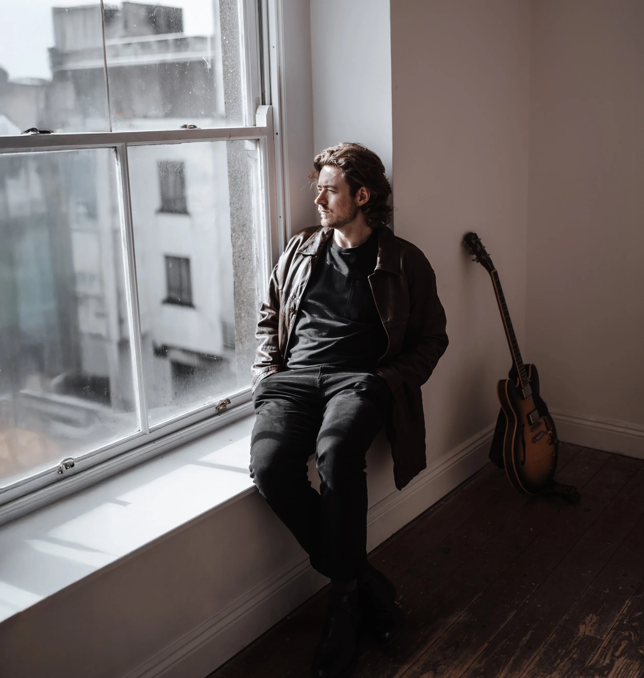 A young man with curly hair sitting on a windowsill, looking out the window with a pensive expression. He is wearing a black shirt and a brown leather jacket. A guitar leans against the wall beside him in a room with dark wooden floors.