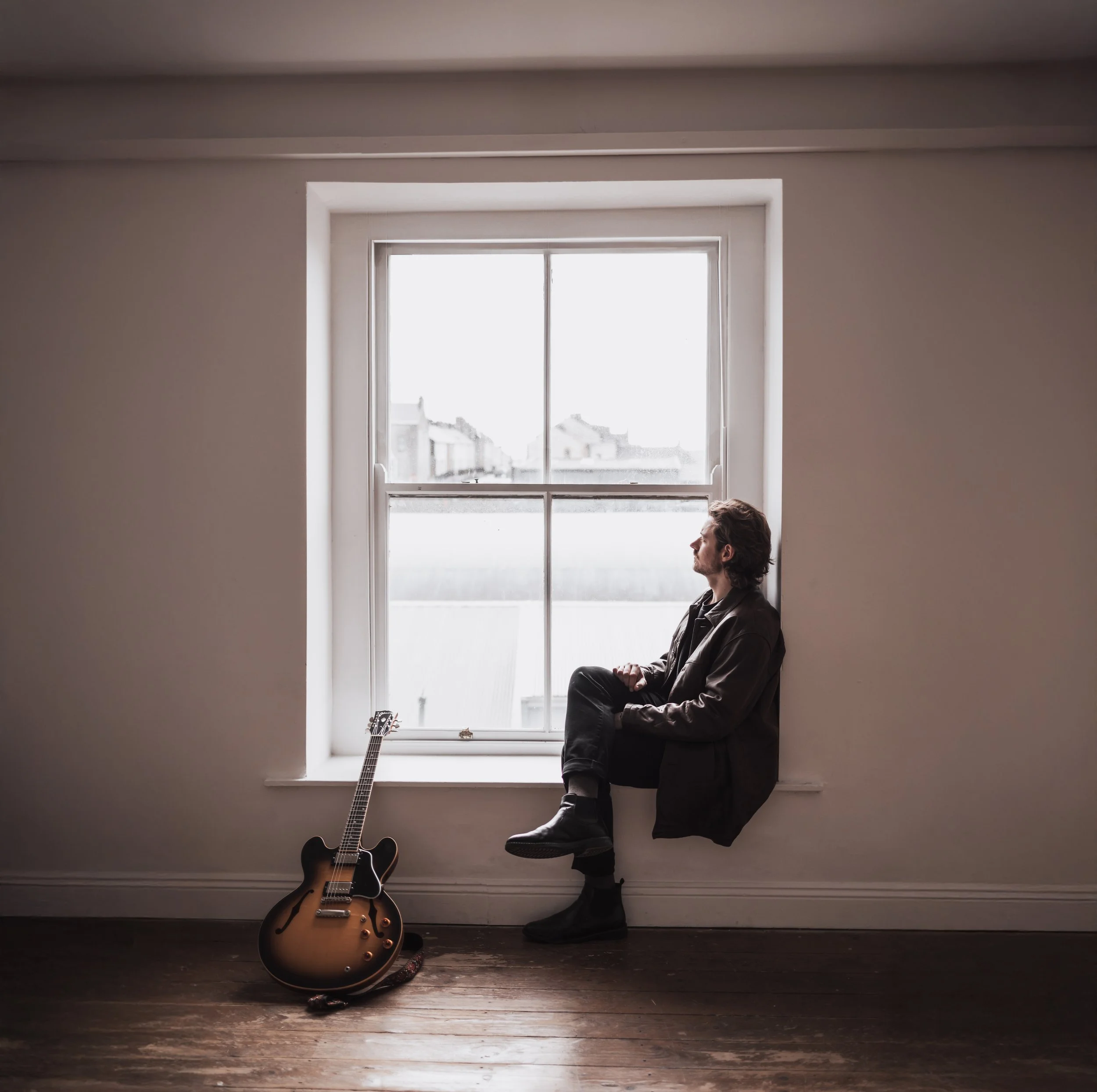 A man sitting on a windowsill looking outside with a guitar leaning against the wall beside him in a minimalist room.
