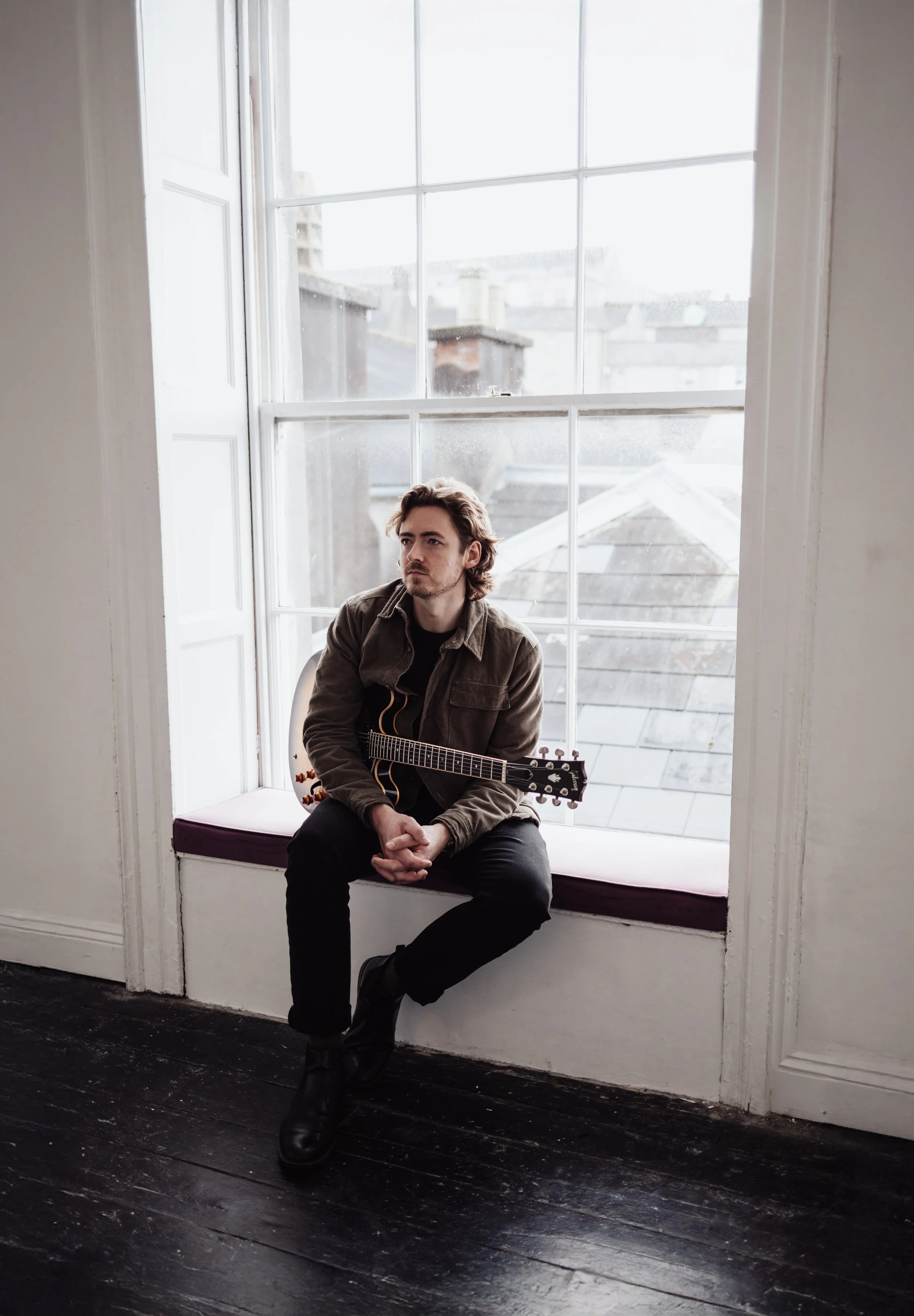 A man sitting on a window seat with a guitar, looking out of a large window in a minimalist room with dark wooden floors.
