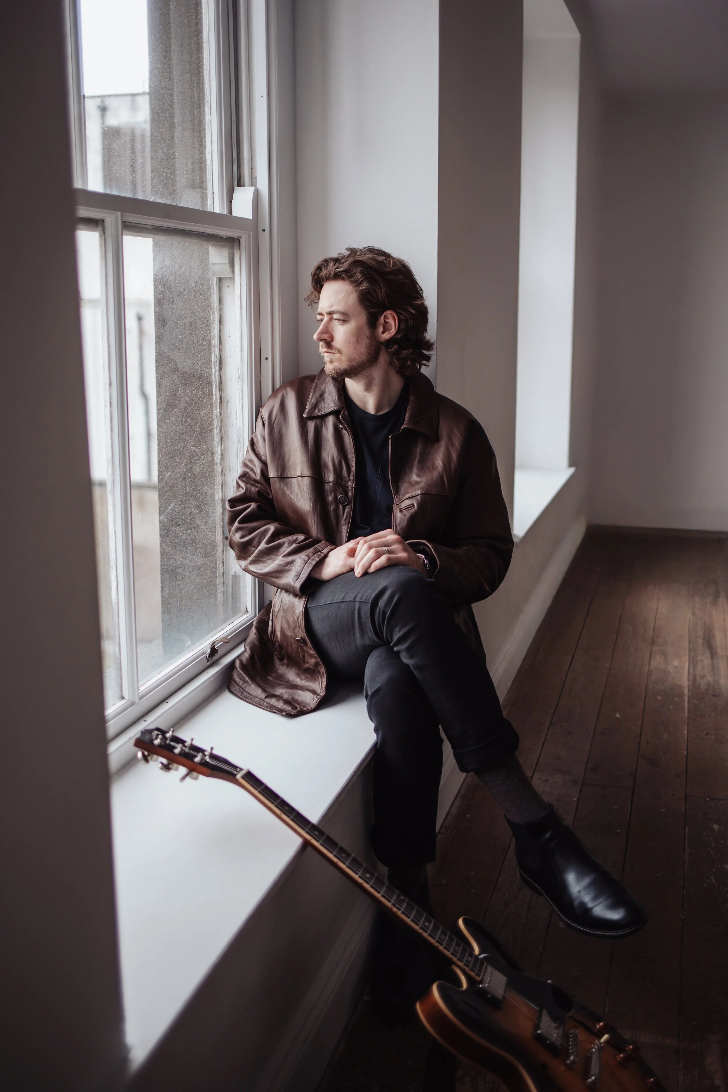 Man sitting on window sill next to an acoustic guitar, looking out a window with sunlight.