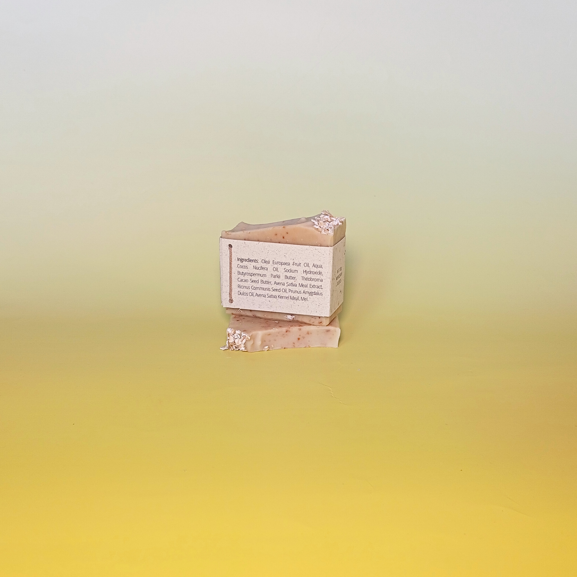 Handmade natural soap bar with a soft beige tone and visible oats, unscented and crafted with milk, honey, and oats, displayed on a pale yellow gradient background wrapped in a grass paper sleeve