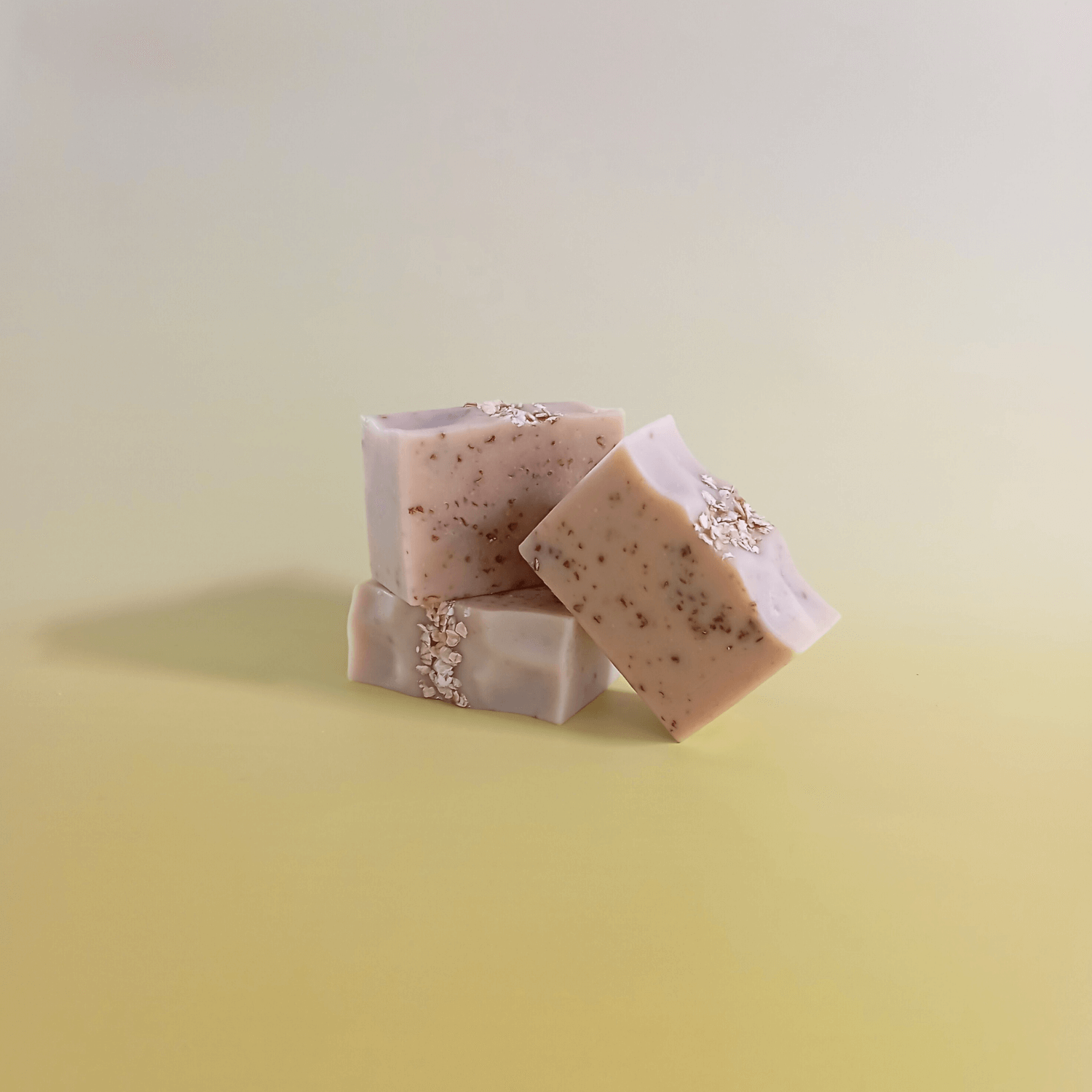 Three handmade unscented natural soap bars with soft beige tones and visible oats, crafted with milk, honey, and oats, artistically stacked on a pale yellow gradient background.