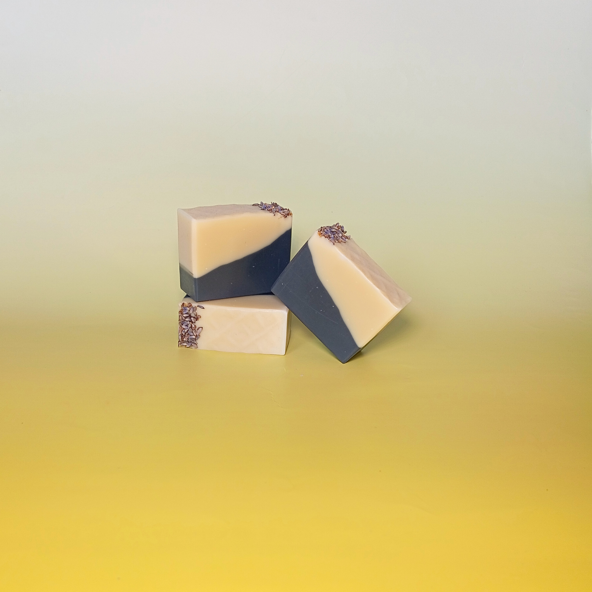 Three handmade soap bars with bold black and cream design, crafted with charcoal powder and scented with orange, patchouli, and lavender essential oils. Stacked on a soft yellow background.