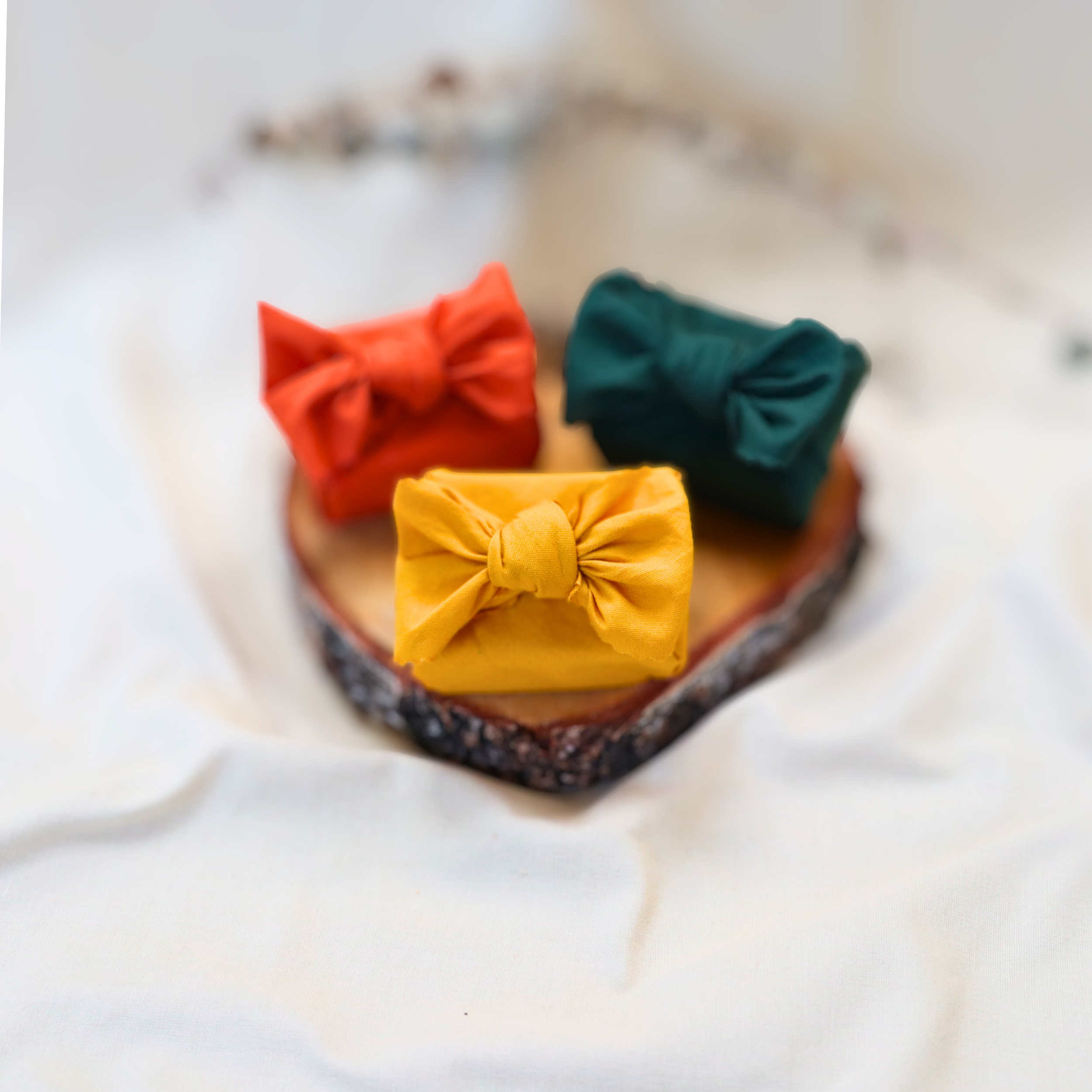 Three small fabric-wrapped gift bundles in mustard yellow, rust orange, and deep green sit on a round wooden slice, arranged in a triangle on soft white fabric with a light, airy background.
