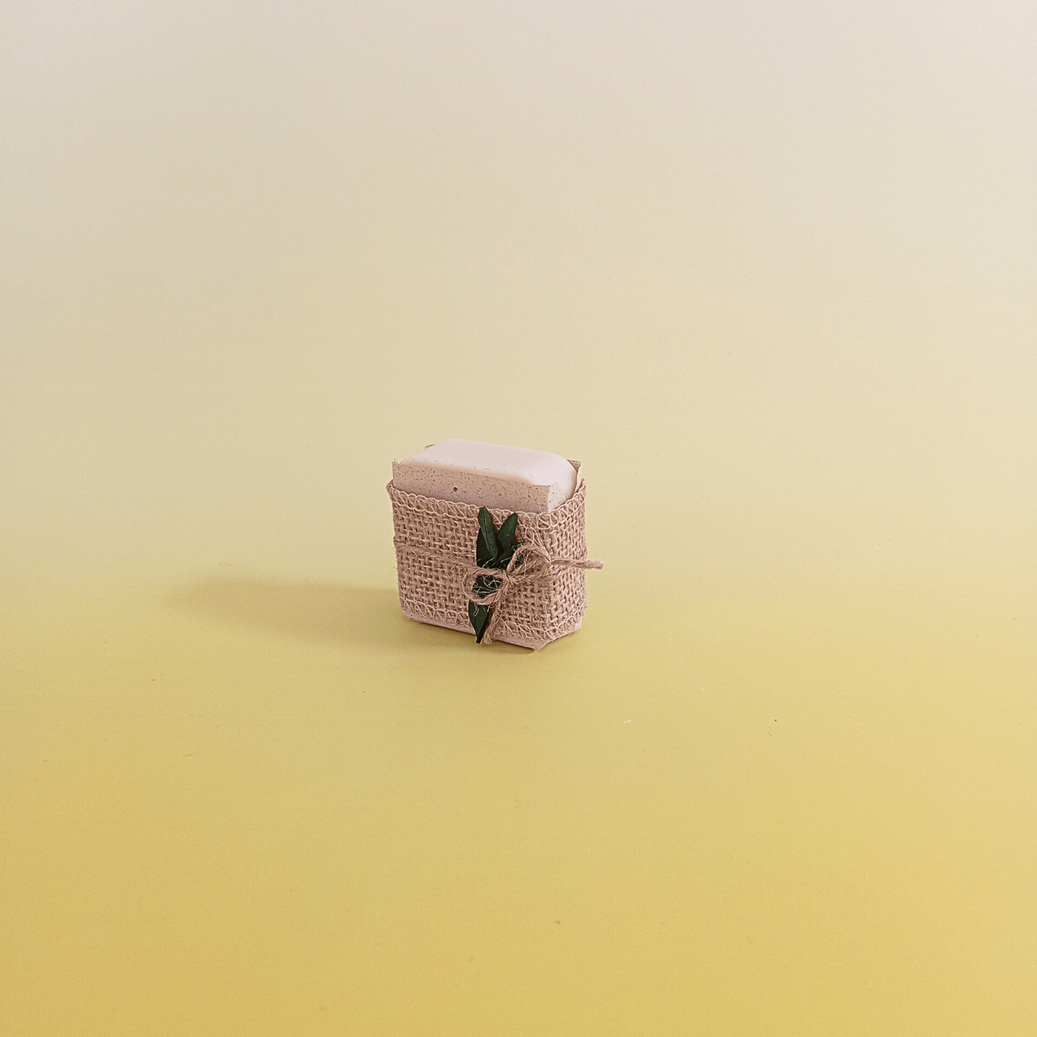 Natural soap bar wrapped in rustic burlap and tied with twine, accented with green botanical detail, displayed on a soft yellow gradient background for a natural and minimal look.