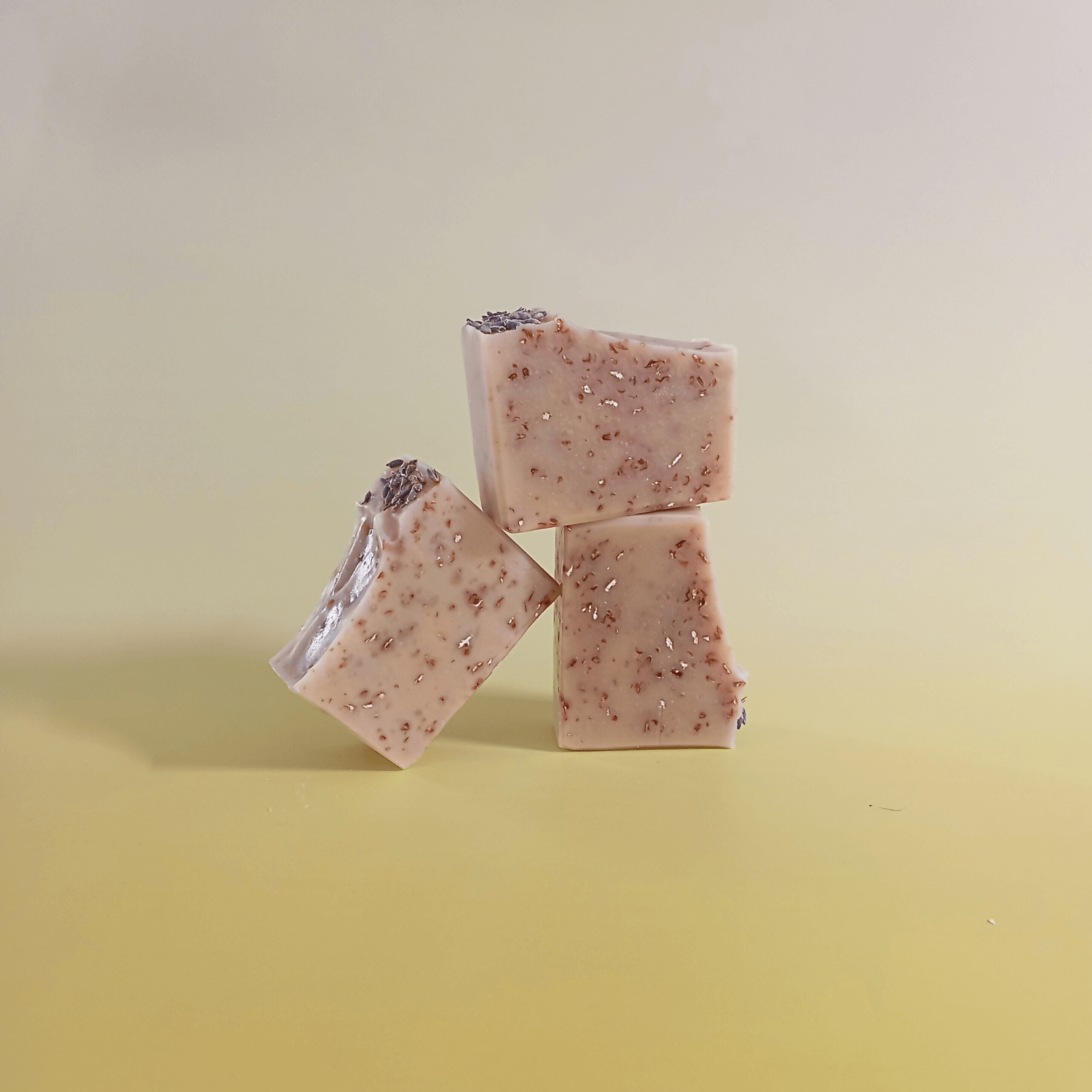 Three handmade natural soap bars with soft beige tones, visible oats and lavender buds, stacked in a minimal composition on a pale yellow gradient background. Scented with milk, honey, and lavender.