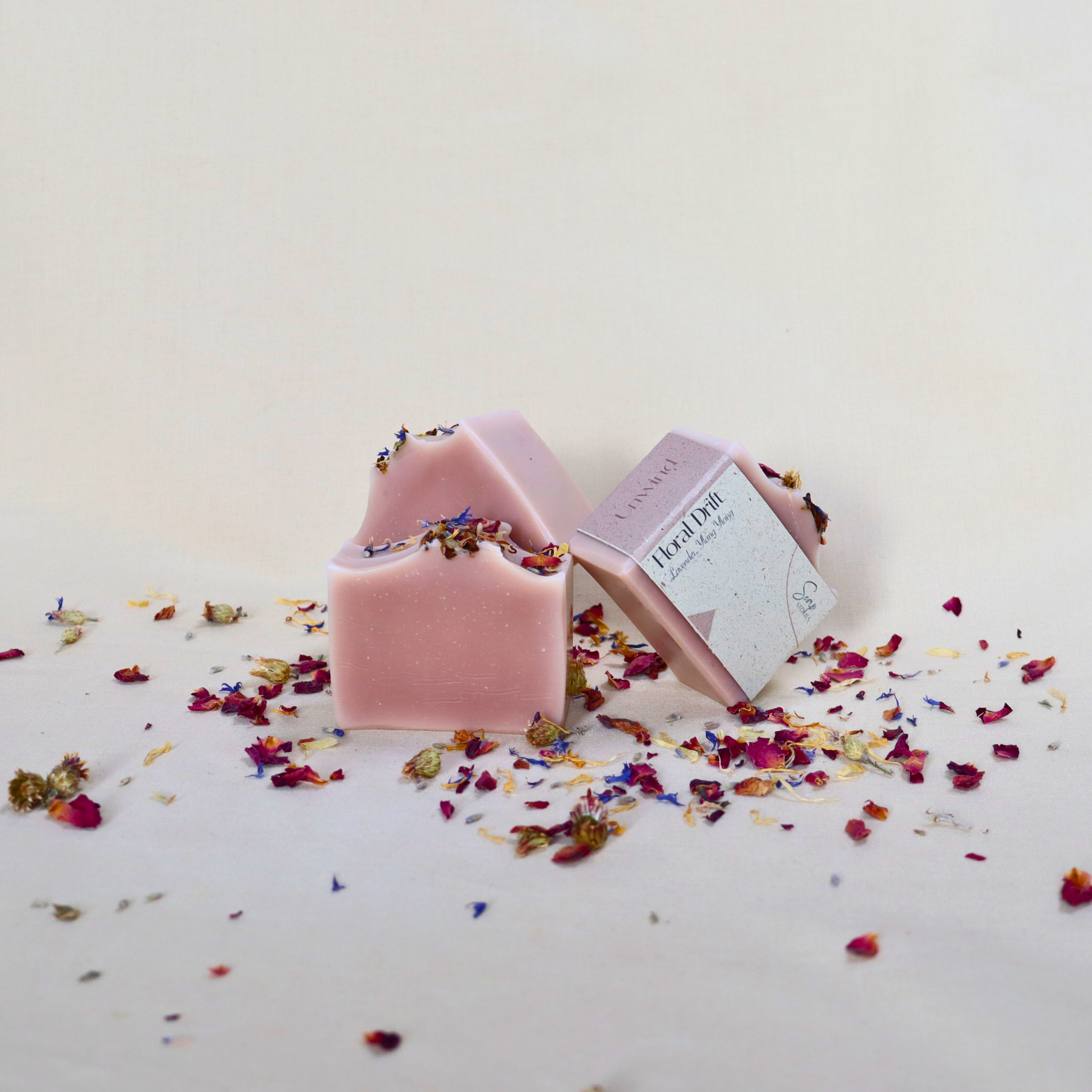 Handmade natural lavender and ylang-ylang soap bar with floral petals on a soft cream fabric background, featuring gentle pink tones and a calming, luxurious look.